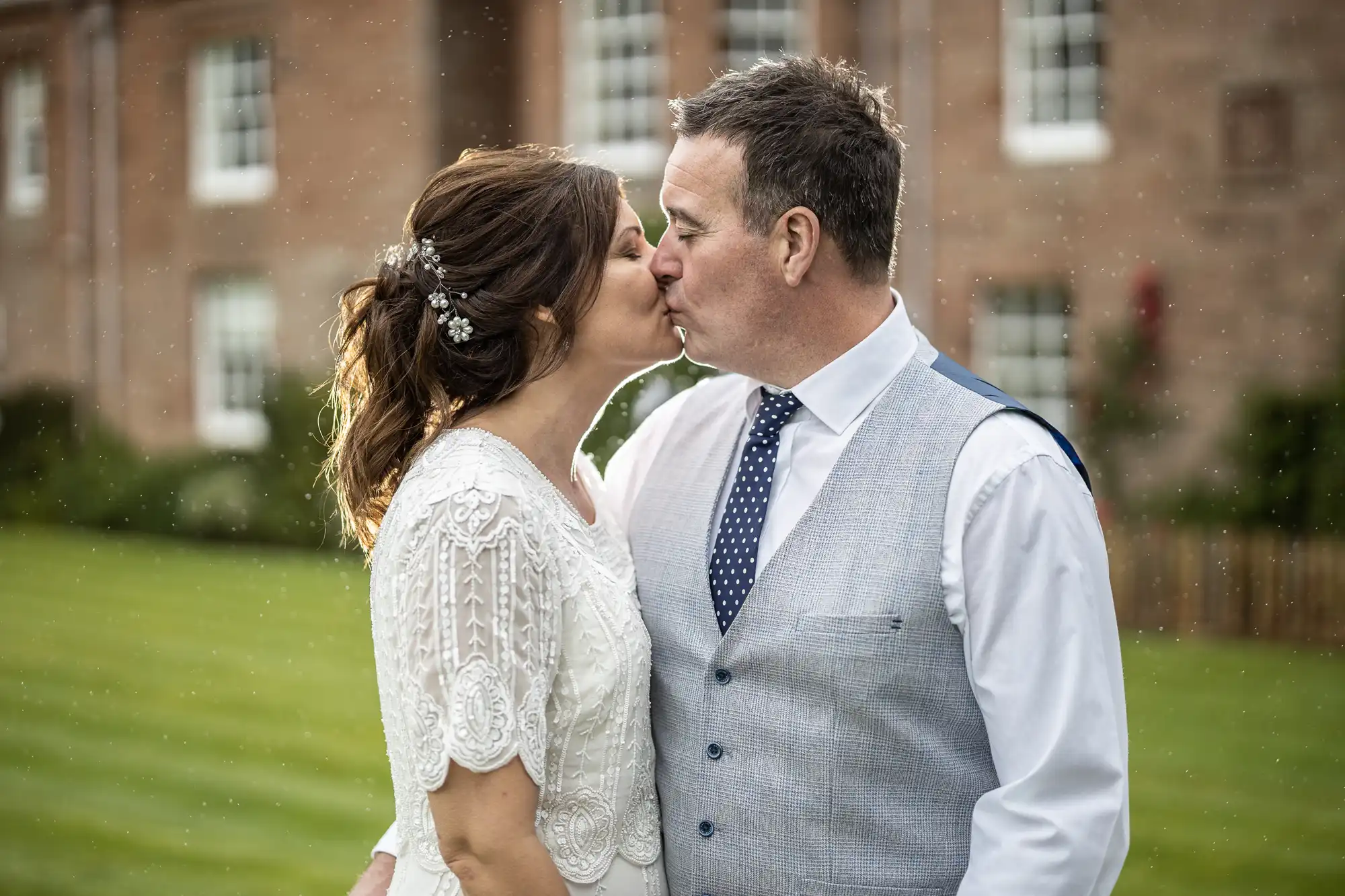 A bride and groom kiss outdoors. The bride wears a white lacy dress with embellished hair, and the groom wears a light grey waistcoat, white shirt, and blue polka-dot tie. They stand on grass with a blurred brick building in the background.