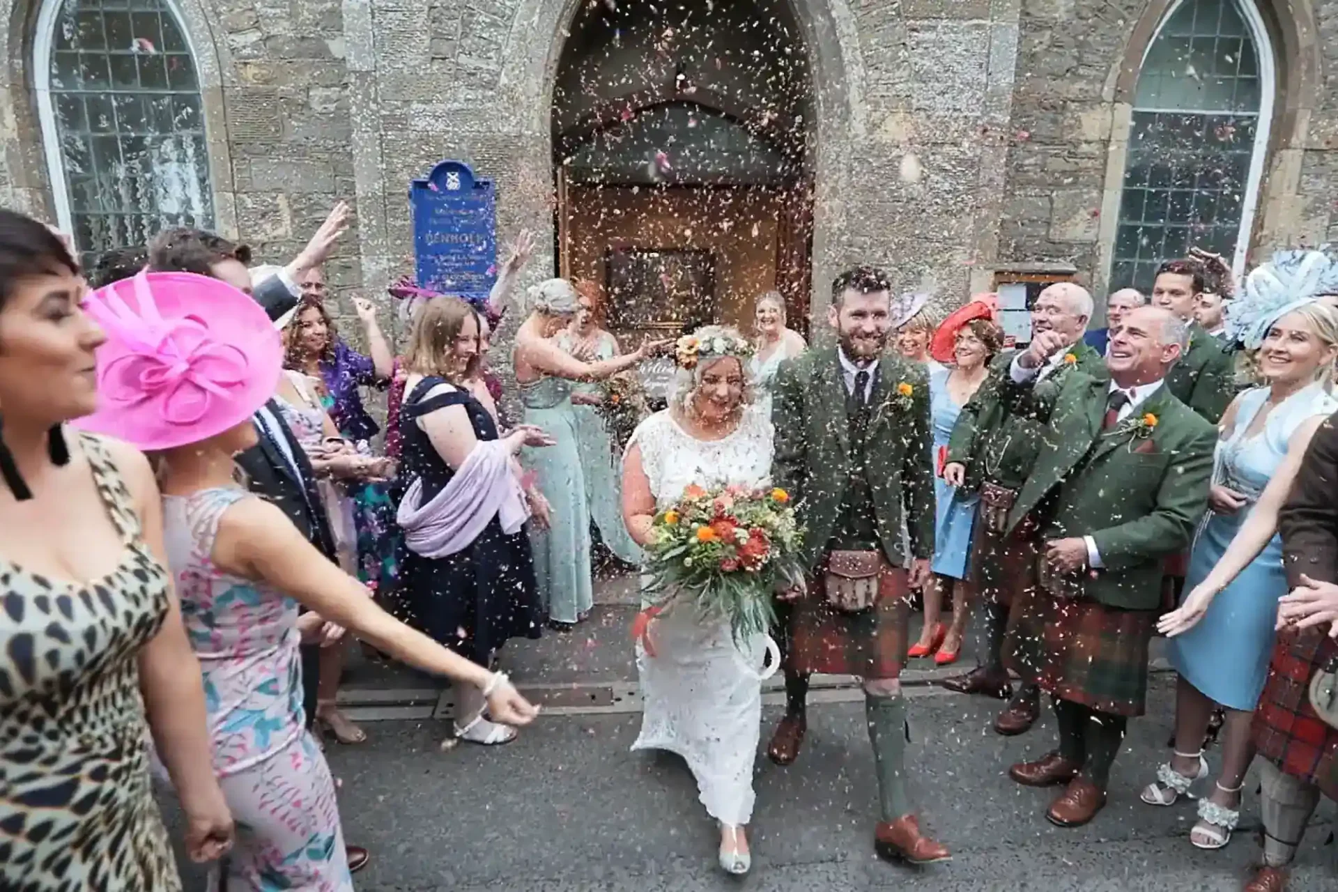 A bride and groom smile as they walk outside Denholm Church, surrounded by joyful guests throwing confetti. The bride wears a white dress and flower crown, holding a bouquet; the groom wears a green suit. Guests celebrate in colourful outfits and hats.