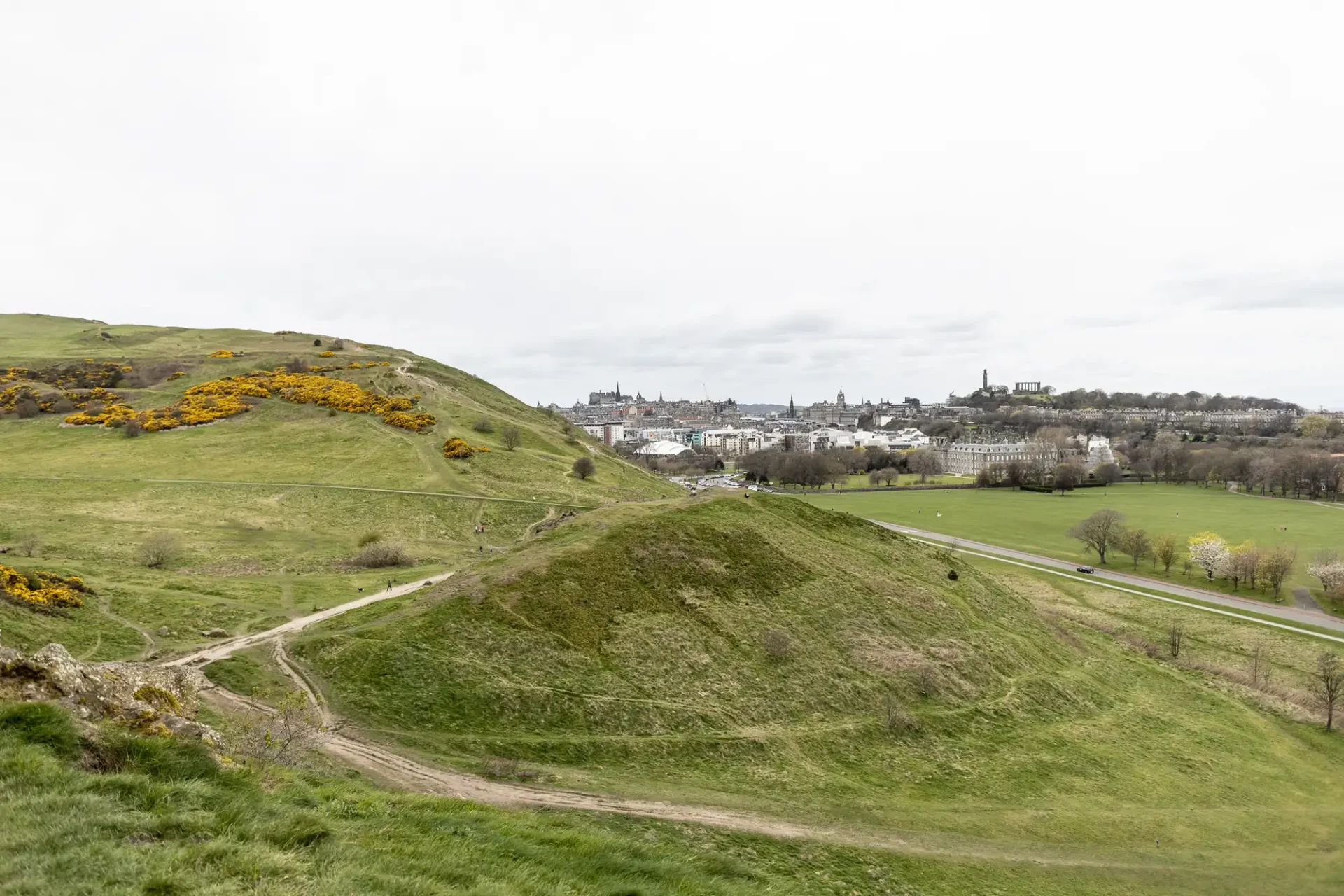 Destination wedding in Edinburgh: A grassy hill with dirt paths overlooks a distant cityscape under a cloudy sky. Sparse trees and a small road are visible in the background.