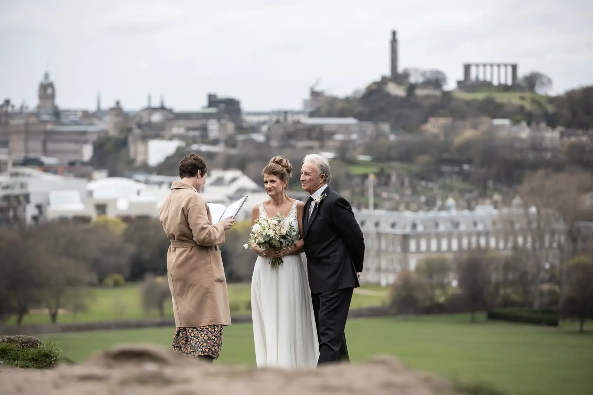 A bride and groom stand together, smiling, as an officiant conducts their wedding ceremony outdoors with a cityscape and greenery in the background.