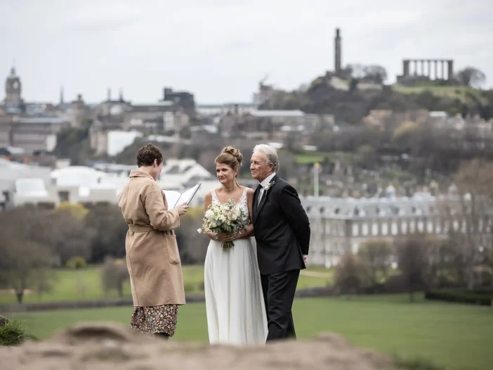 A bride and groom stand together, smiling, as an officiant conducts their wedding ceremony outdoors with a cityscape and greenery in the background.