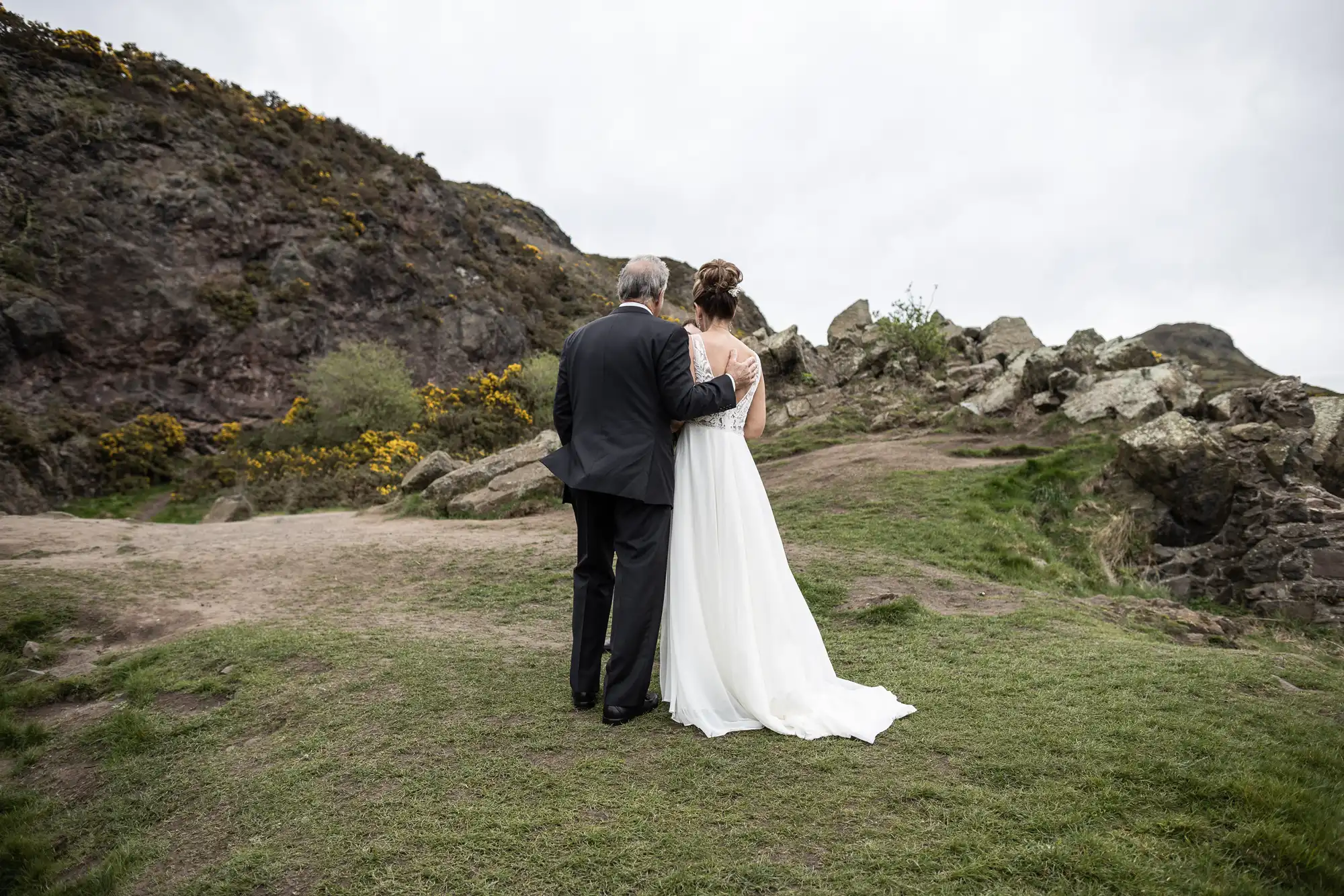 A couple dressed in formal attire stands side by side on a grassy, rocky landscape, facing away from the camera and looking at the mountainous scenery.