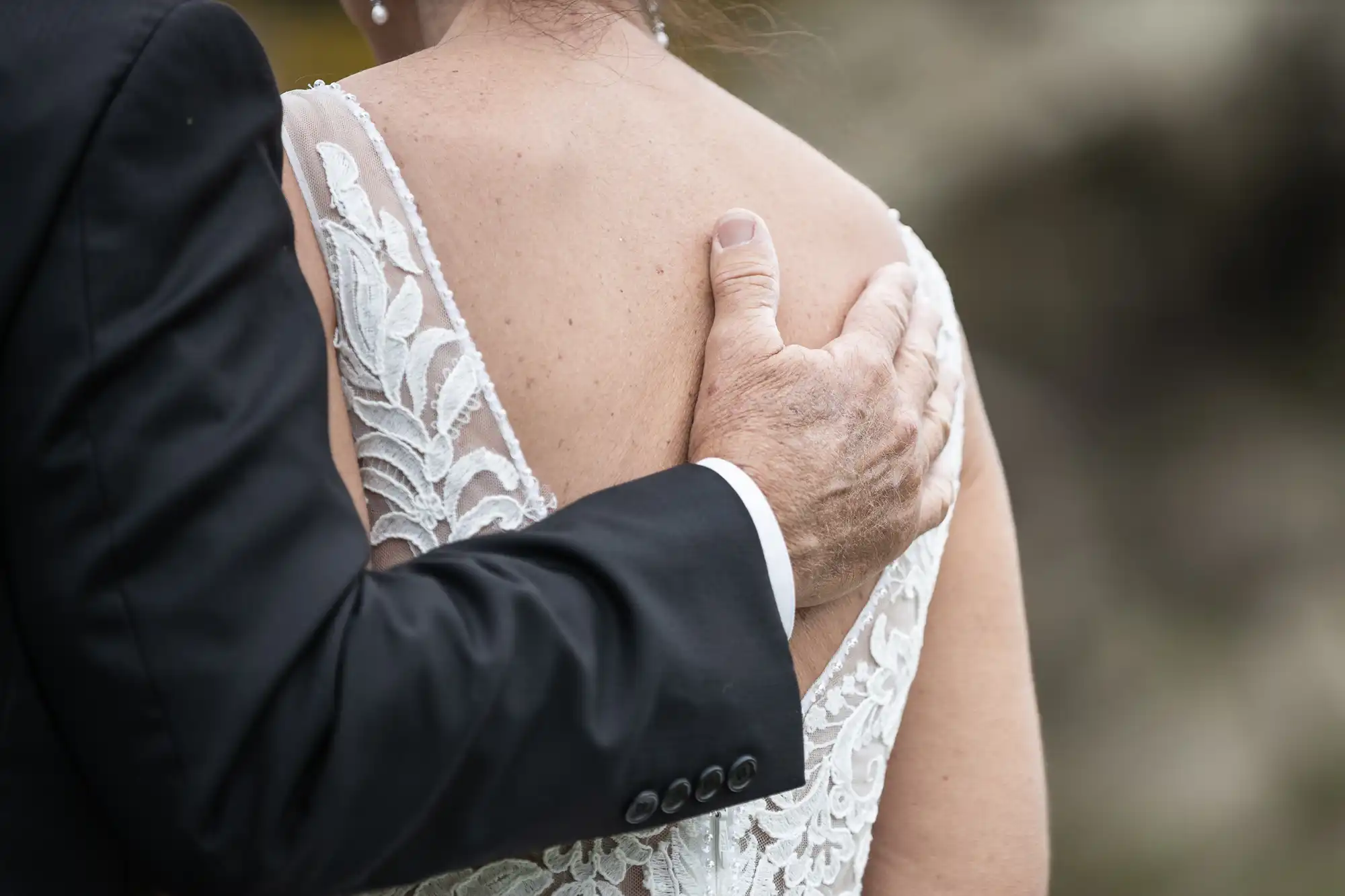 A person in a black suit gently places their hand on the back of a person wearing a white lace garment.