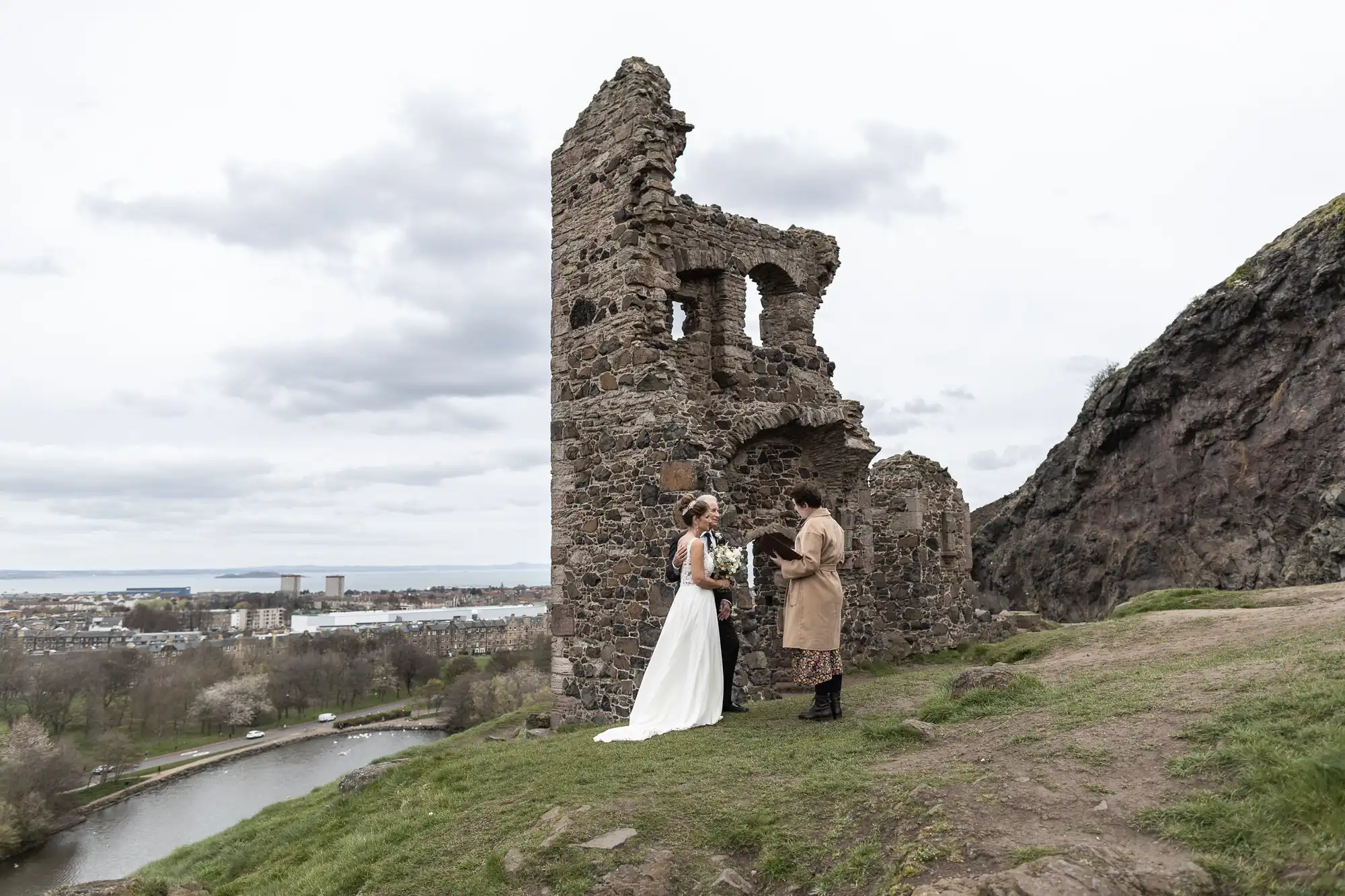 A couple stands in wedding attire near a historic stone ruin on a grassy hilltop overlooking a body of water and distant cityscape.