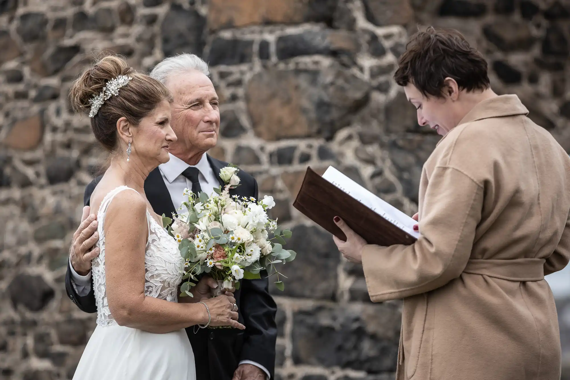 An elderly couple, dressed in wedding attire, stands in front of a stone wall. The bride holds a bouquet, and an officiant, holding an open book, reads to them during their wedding ceremony.