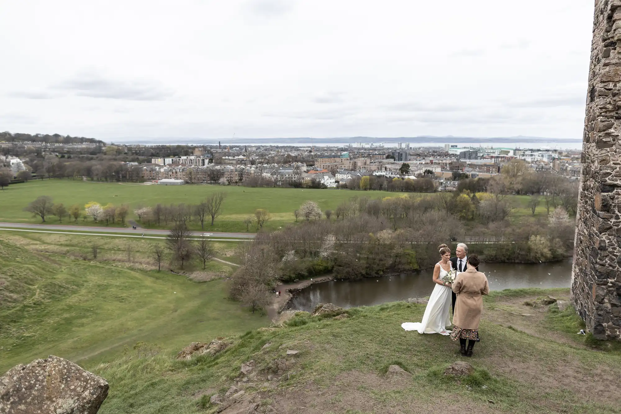 A couple in wedding attire stands with an officiant on a cliffside, overlooking a town, river, and fields. The scene includes a cloudy sky and a historic stone structure.