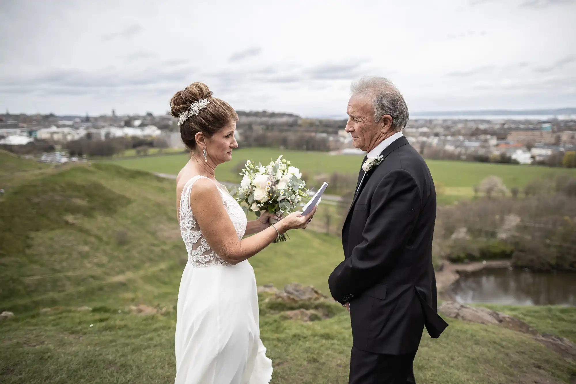 A bride and groom stand outdoors on a grassy hill with the bride holding a bouquet and reading vows to the groom. The background shows a cloudy sky and distant landscape with buildings and greenery.