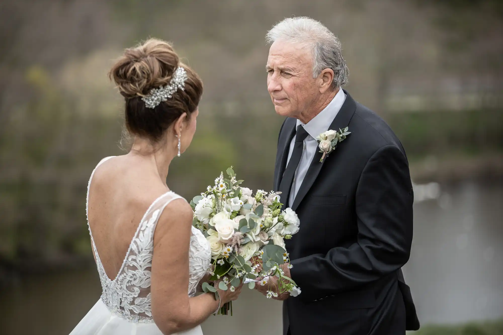 A bride and an older man in a suit, possibly the father of the bride, are standing outside. The bride is holding a bouquet, and they are looking at each other. The background is blurred greenery.