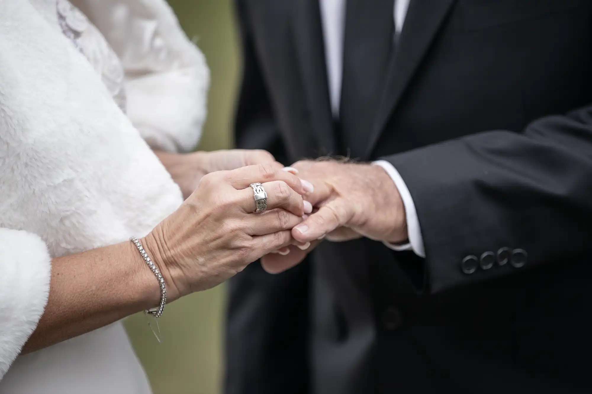 A couple in formal attire holds hands, with the woman's focus on placing a ring on the man's finger.