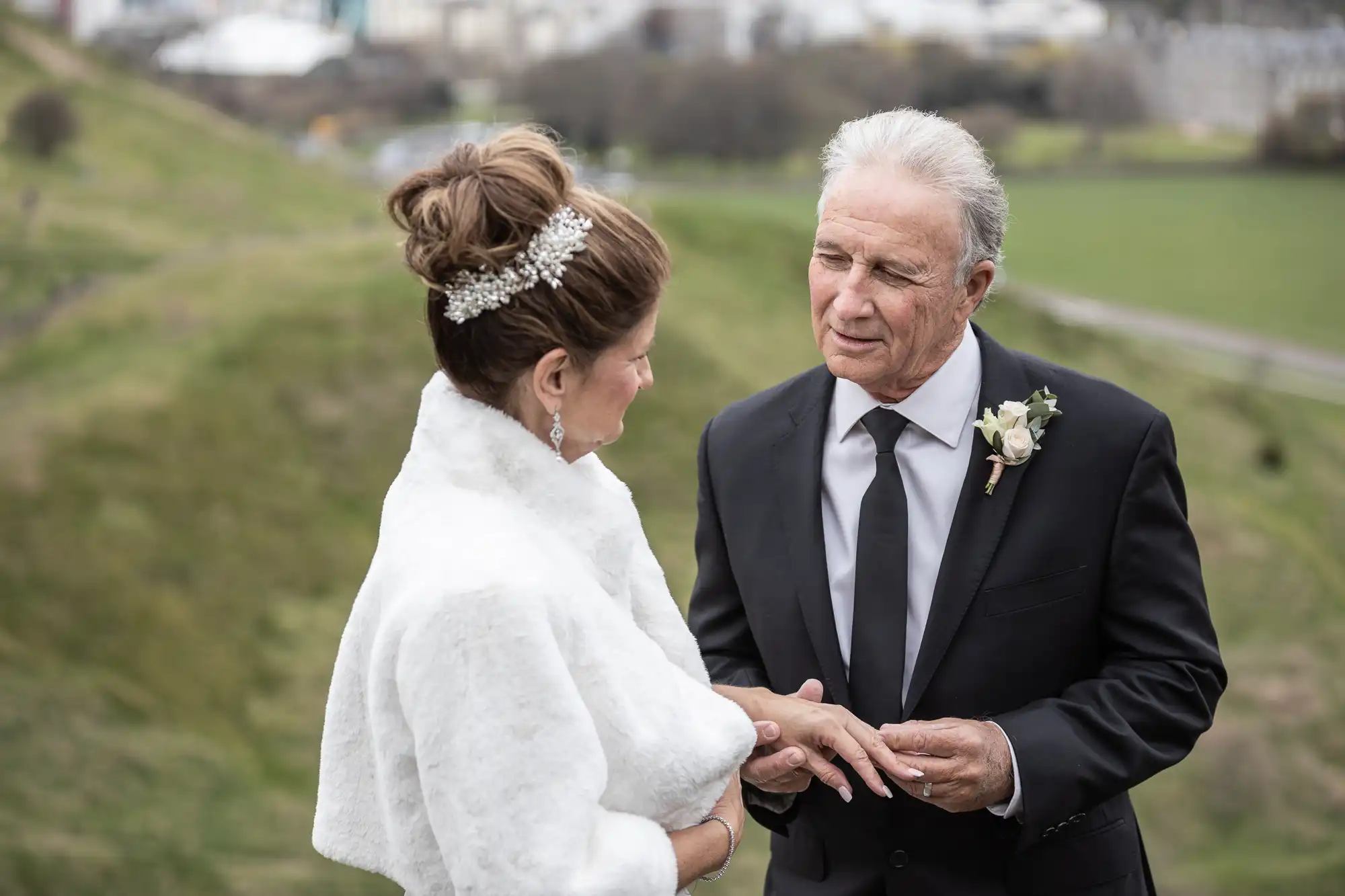 An elderly couple dressed in formal attire stand outside. The man, wearing a black suit, holds the woman's hand and looks at her. The woman, clad in a white fur shawl and adorned with a hair accessory, gazes back.