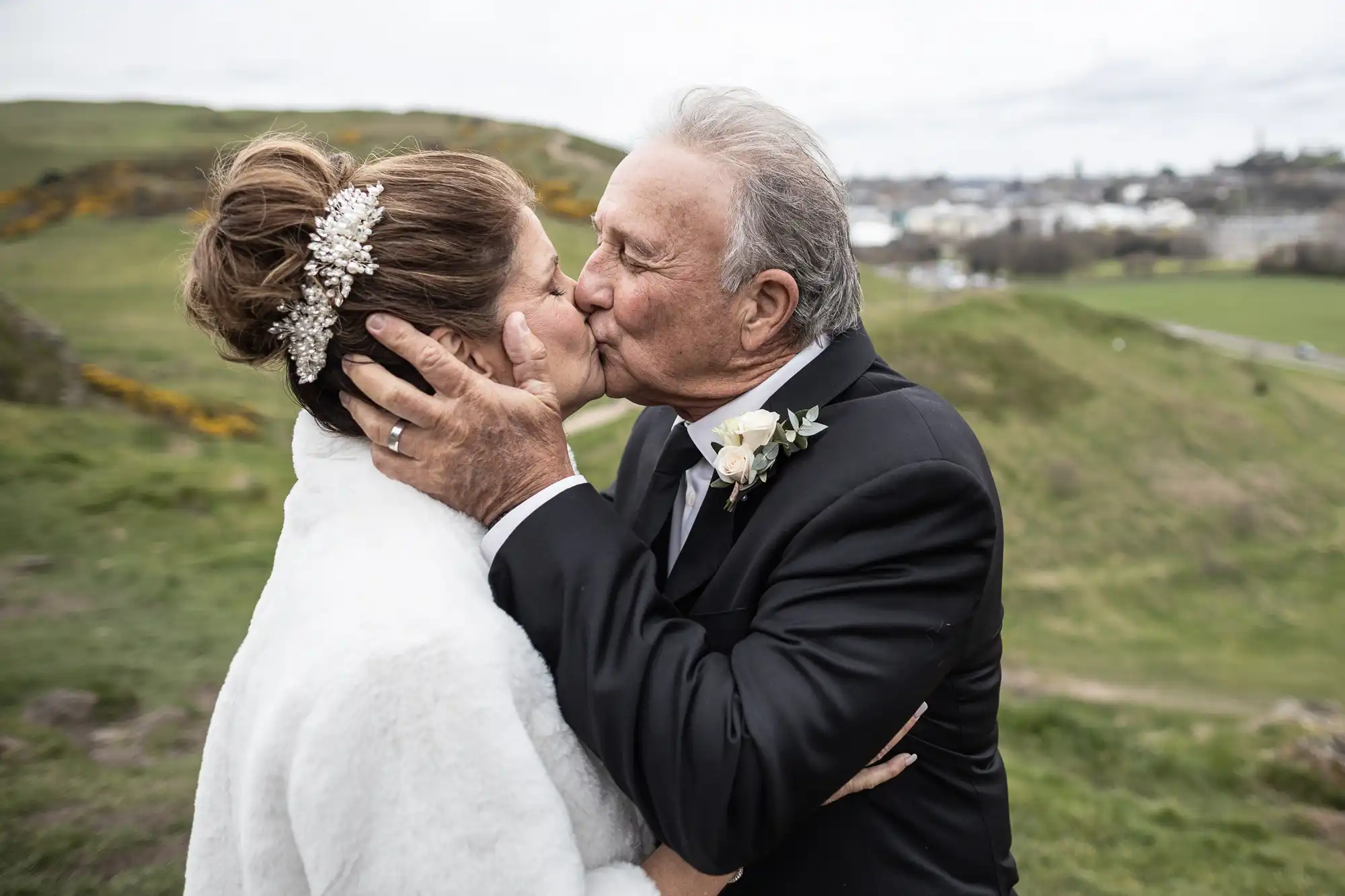 An elderly couple, dressed in wedding attire, share a kiss outdoors in a hilly landscape. The woman wears a white fur shawl and decorative hairpiece, while the man wears a black suit with a boutonnière.