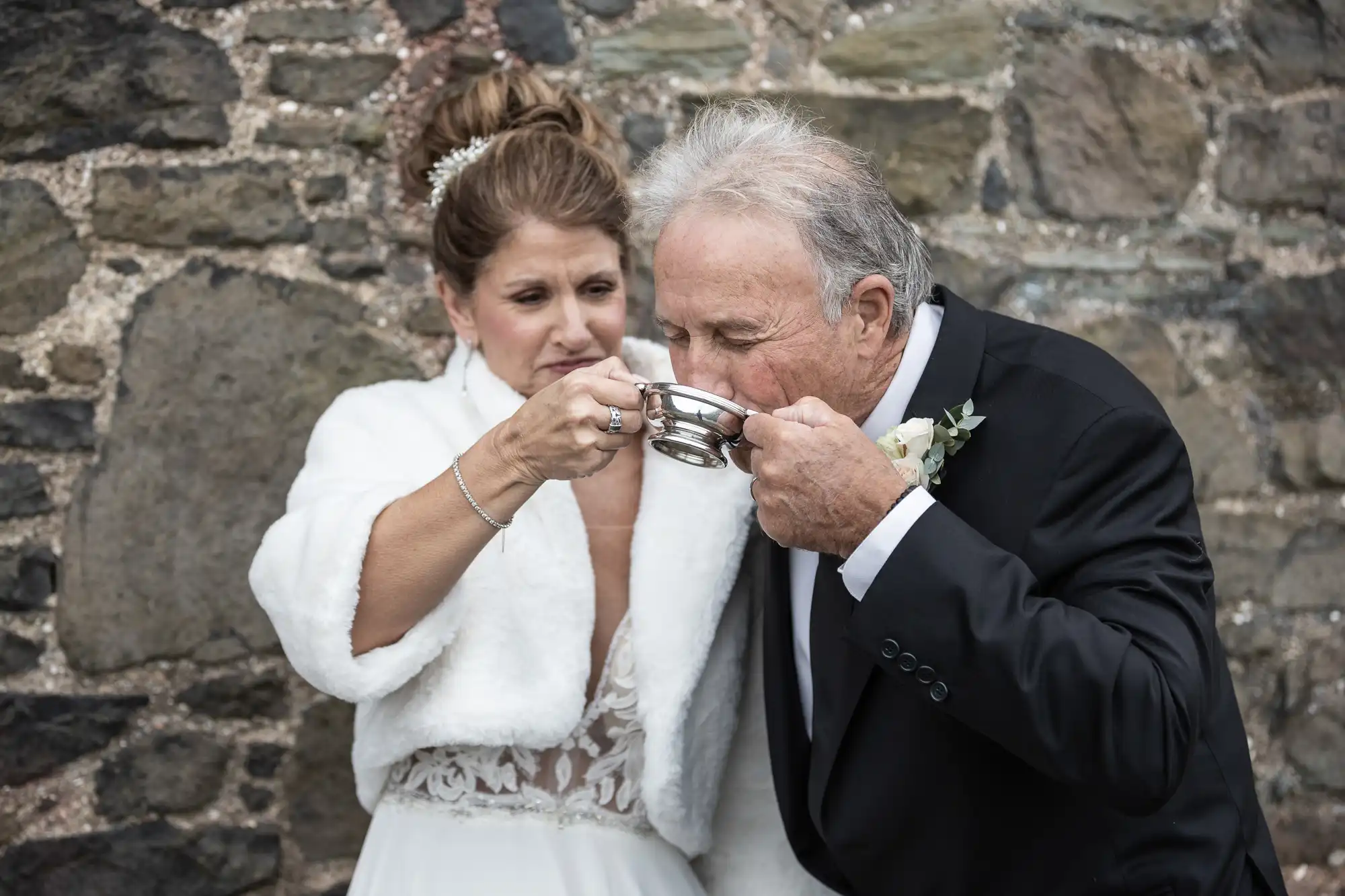 A bride in a white dress and fur stole helps an older man in a black suit drink from a silver cup against a stone wall backdrop.