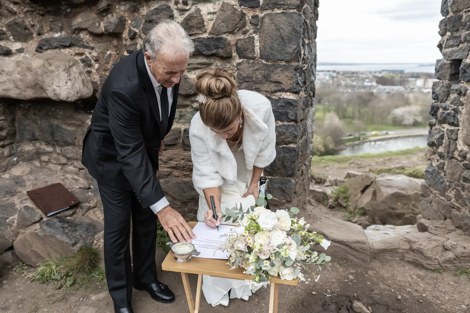 A man in a suit and a woman in a white outfit sign documents on a small table near a stone wall. A bouquet of white flowers is on the table. The background shows a scenic outdoor landscape.