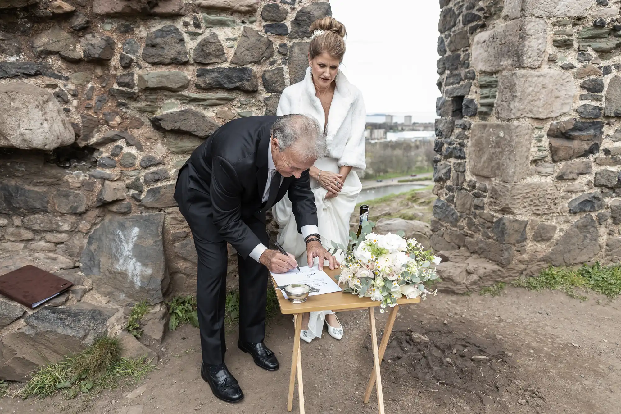 A groom in a black suit signs a document on a small wooden table while the bride in a white dress and shawl stands beside him. They are outdoors next to a stone wall with a bouquet on the table.