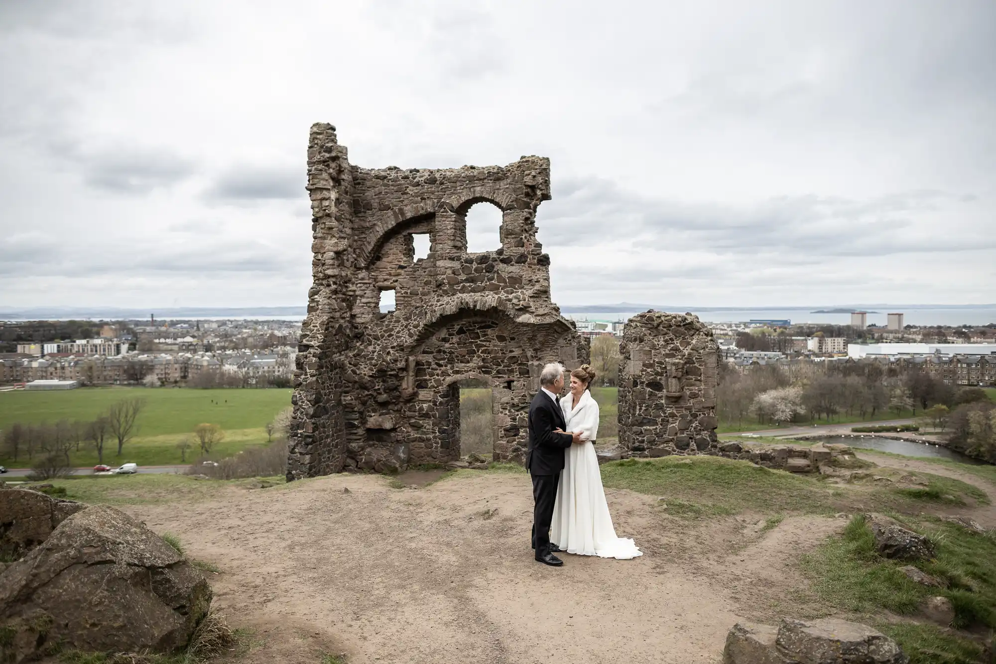 A bride and groom stand in front of a historic stone ruin on a hilltop with a sprawling cityscape and park in the background.