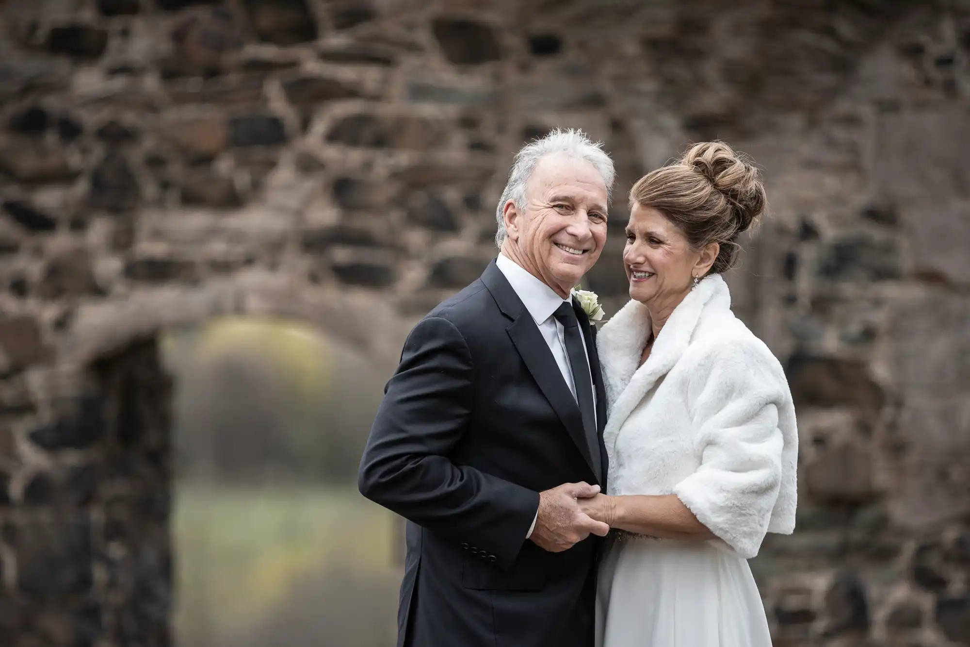 An older couple dressed in formal attire, with the man in a suit and the woman in a white dress with a fur stole, pose together smiling in front of a stone archway.