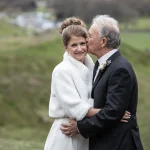 An elderly couple in wedding attire embraces outdoors. The woman smiles while the man kisses her on the cheek. They appear to be at a countryside location with green fields in the background.