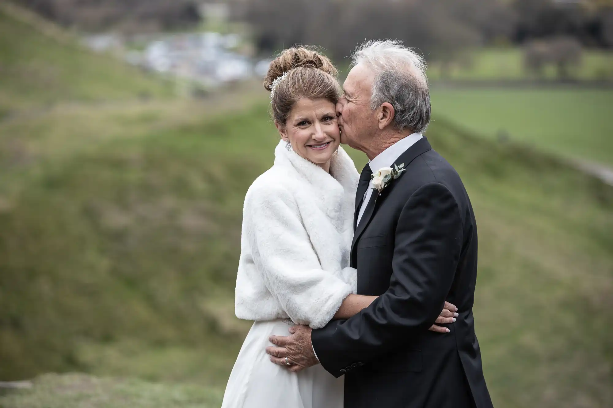 An elderly couple in wedding attire embraces outdoors. The woman smiles while the man kisses her on the cheek. They appear to be at a countryside location with green fields in the background.