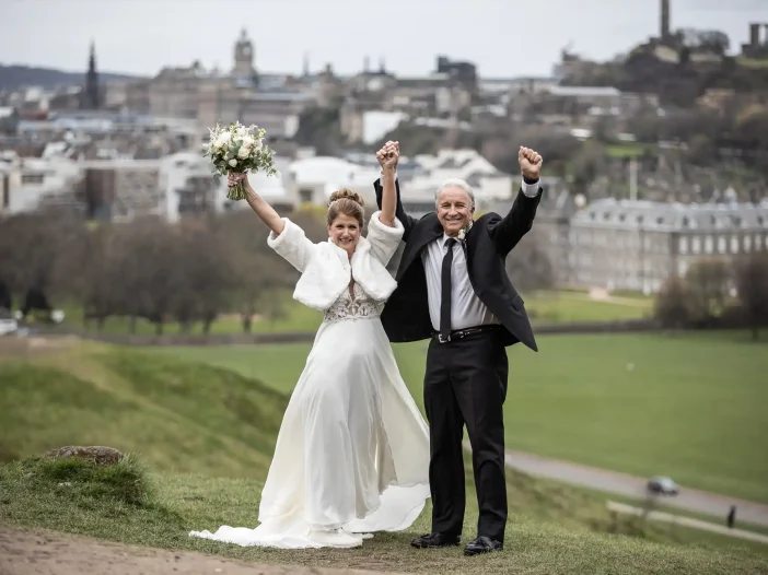 destination wedding in Edinburgh Bride and groom standing on a grassy hill with arms raised in celebration, cityscape in the background.