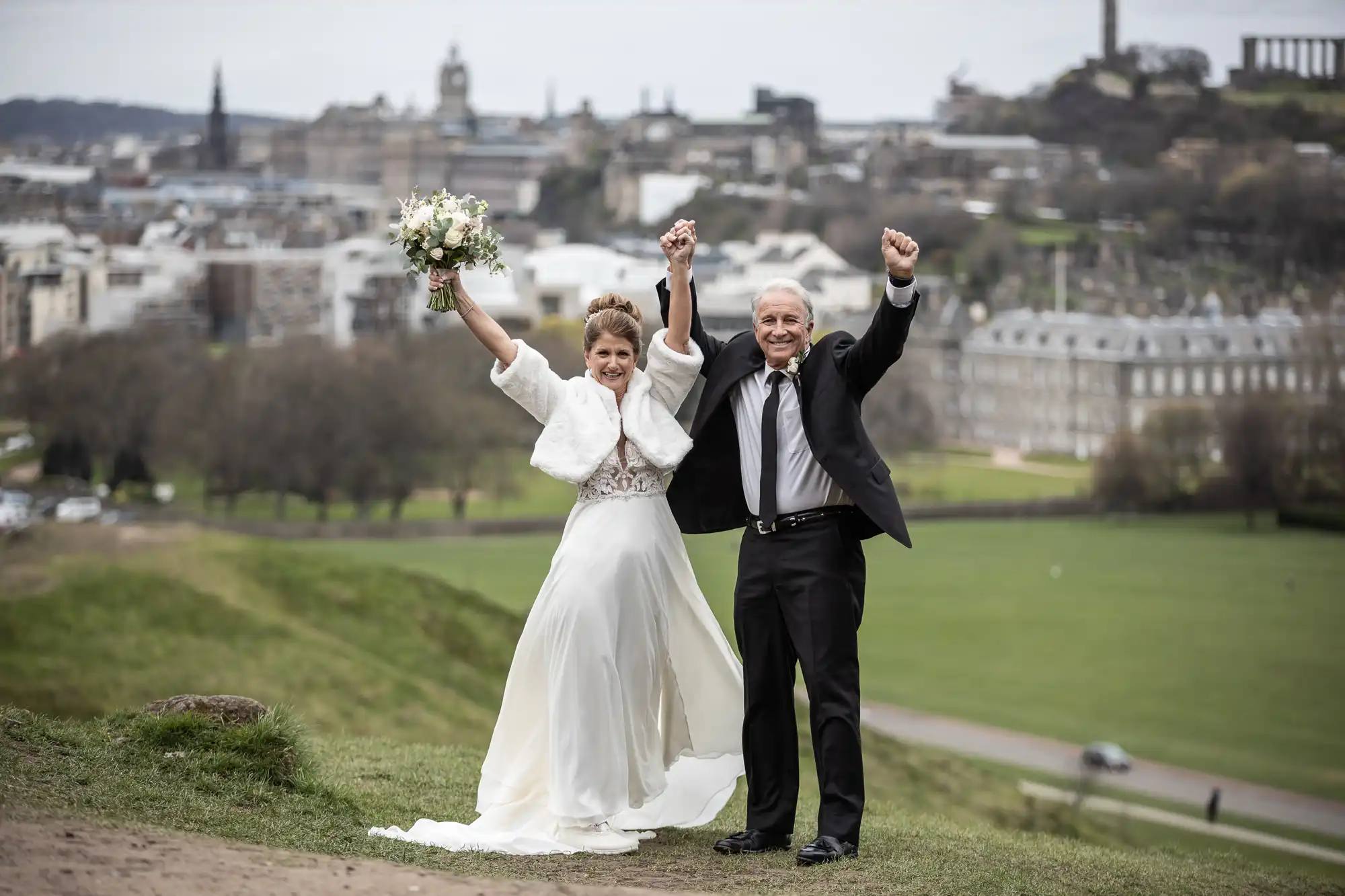 destination wedding in Edinburgh Bride and groom standing on a grassy hill with arms raised in celebration, cityscape in the background.