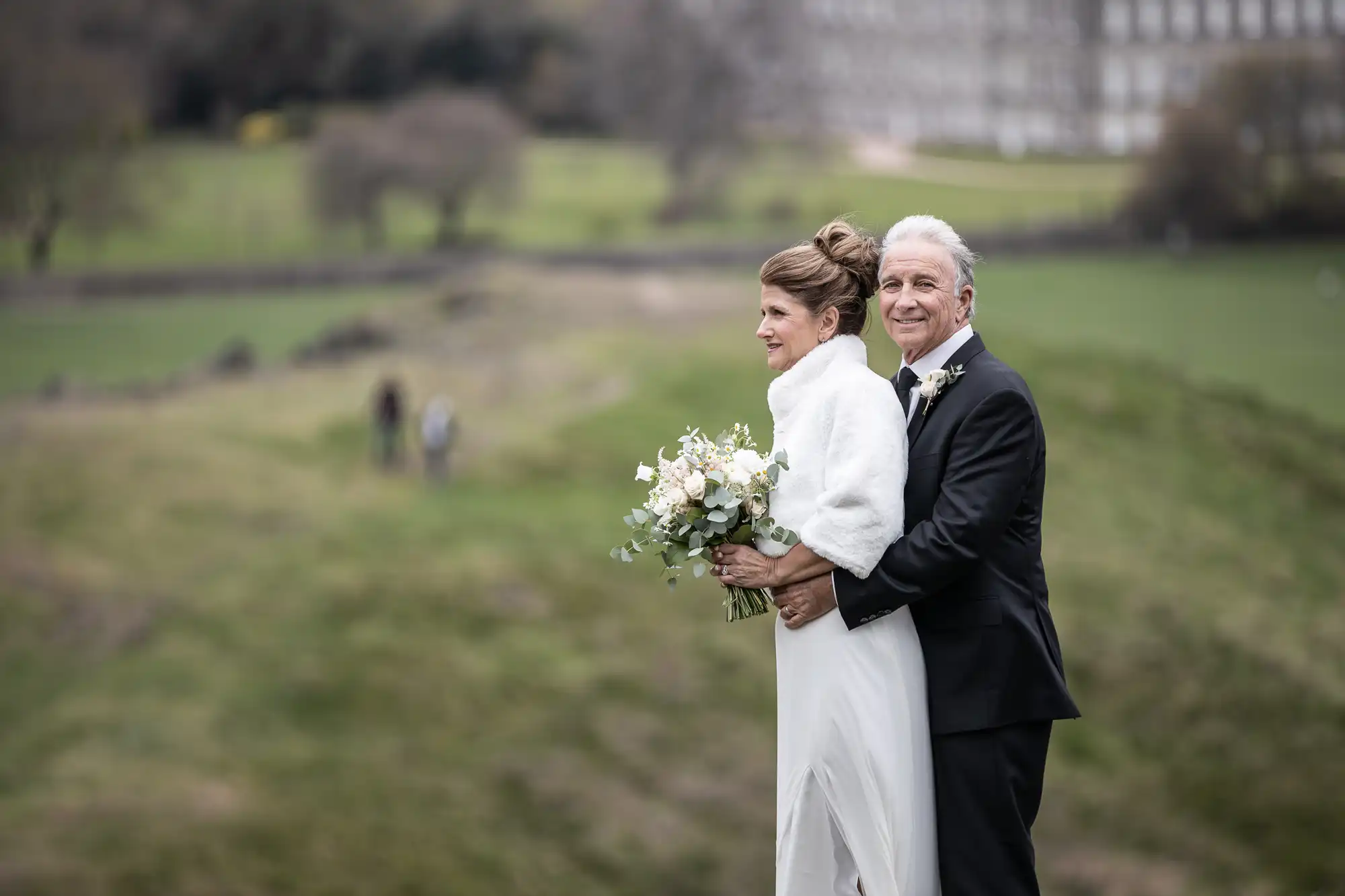 A bride and groom stand together outdoors, the groom hugging the bride from behind. The bride holds a bouquet, and both are dressed in formal attire against a scenic green landscape background.