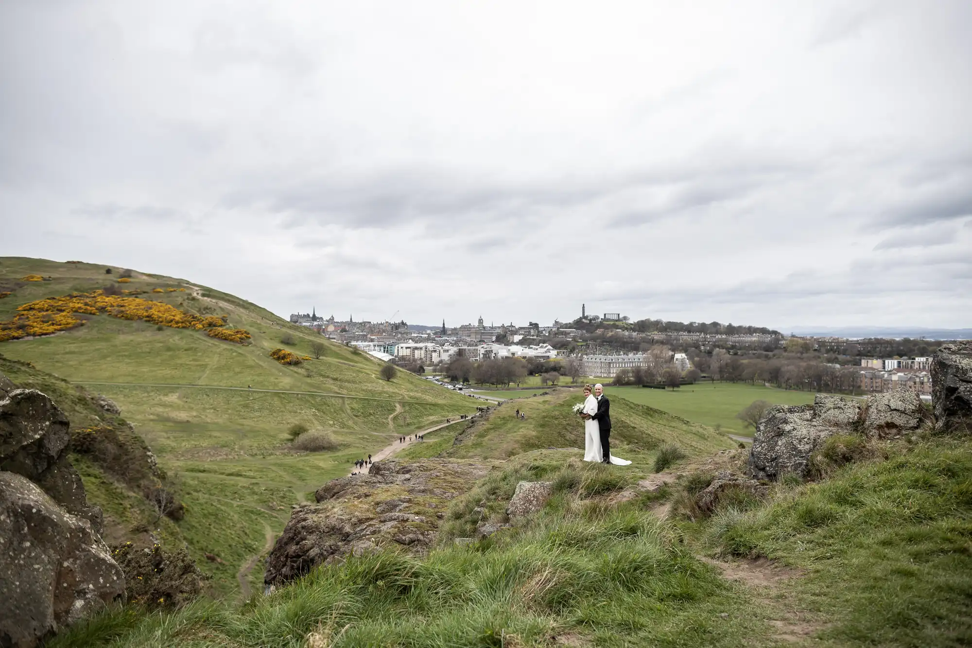 A couple wearing a tuxedo and a wedding dress stand on a rocky outcrop overlooking a green hilly landscape with a city in the background.