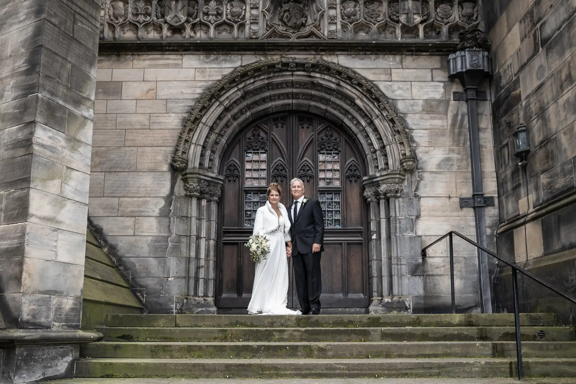 A couple stands in wedding attire on the steps of a historic stone building with an ornate arched wooden door in the background. The bride holds a bouquet and wears a white dress, the groom a black suit.