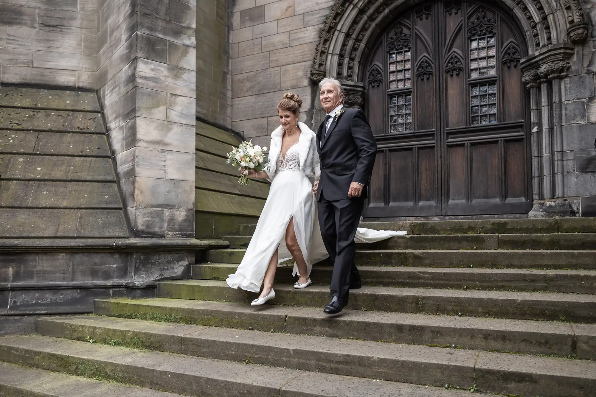 A bride in a white dress holding a bouquet walks down stone steps beside an older man in a black suit, outside a gothic-style building.