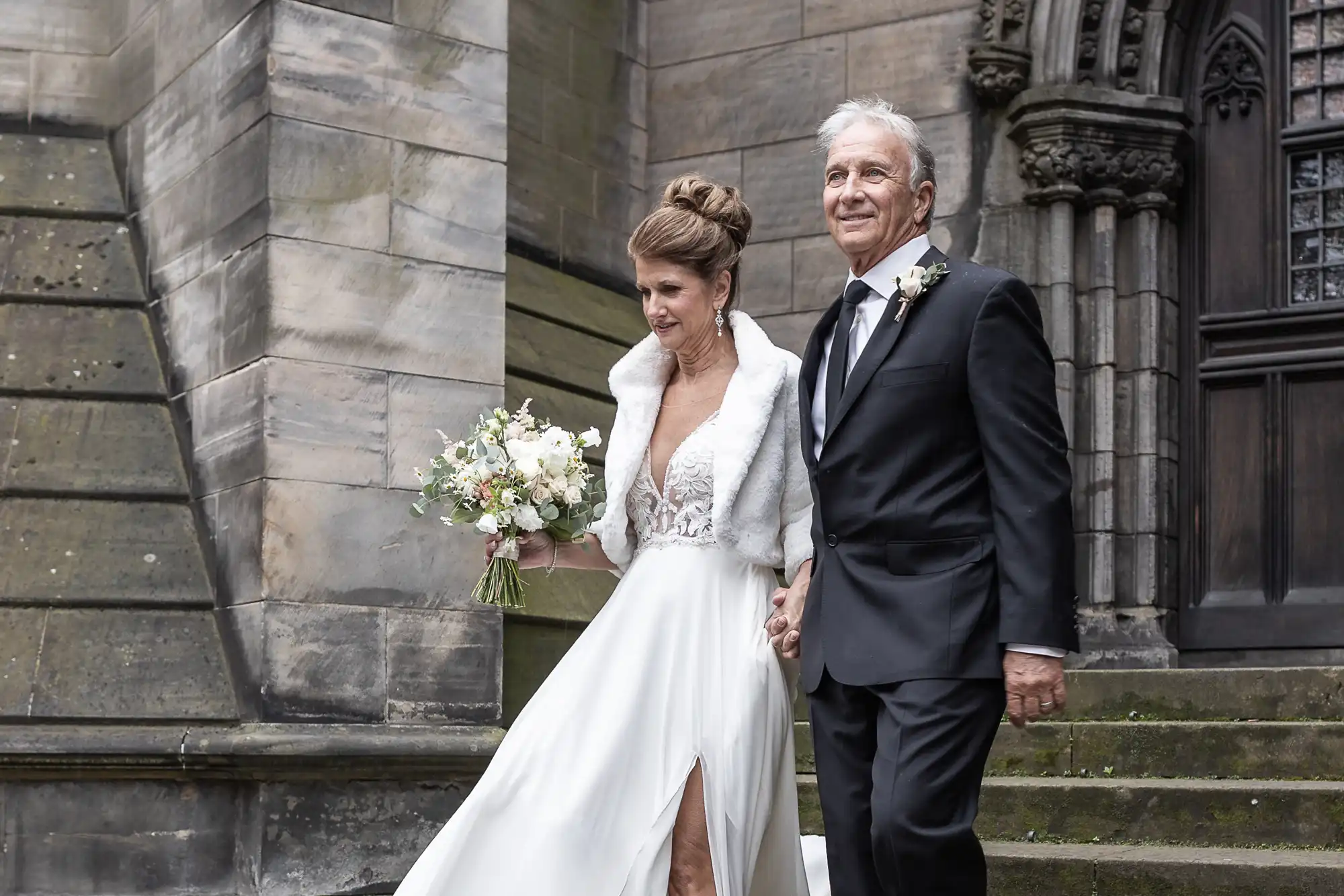 A bride in a white gown and a groom in a black suit hold hands while descending stone steps outside a building.