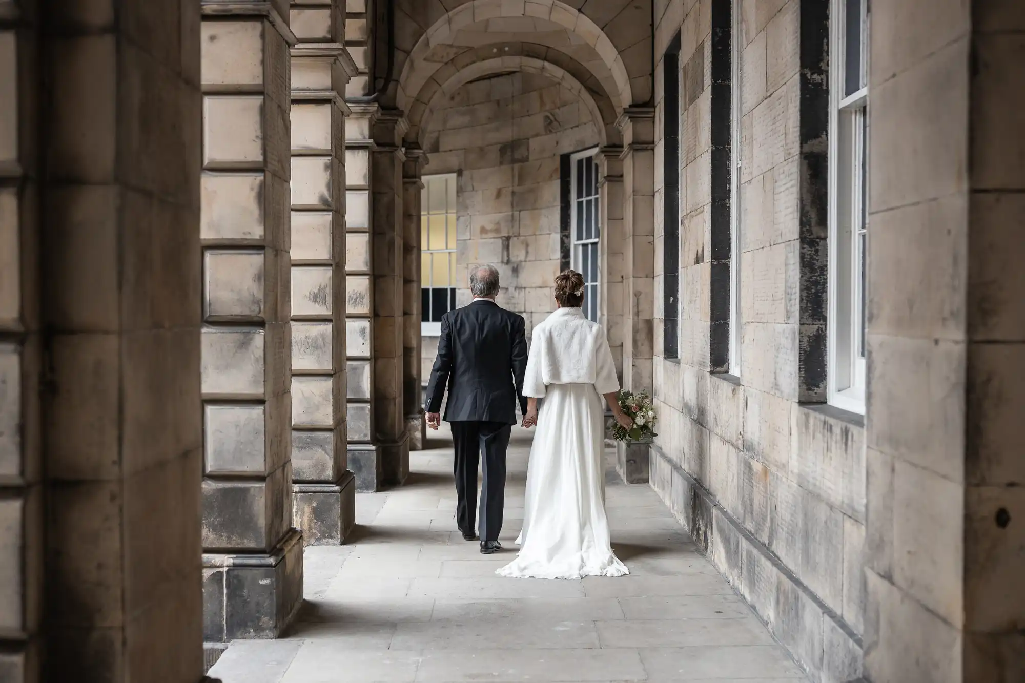 A bride and groom are walking hand in hand down a stone corridor with arches, both dressed in formal attire.