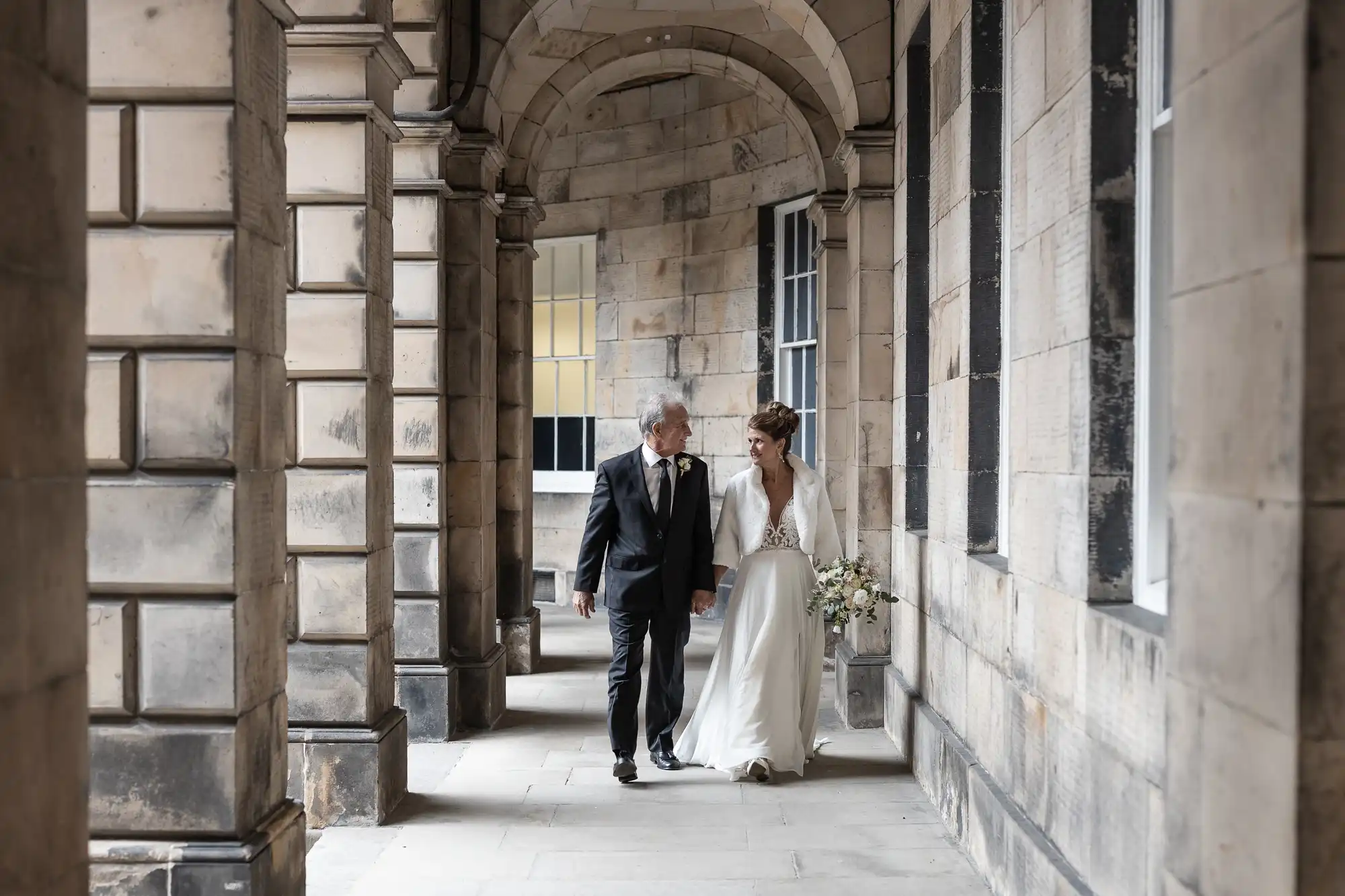 A bride and groom walk hand in hand through a stone archway hallway, both dressed in formal wedding attire. The groom wears a black suit and tie, while the bride wears a white dress and carries a bouquet.