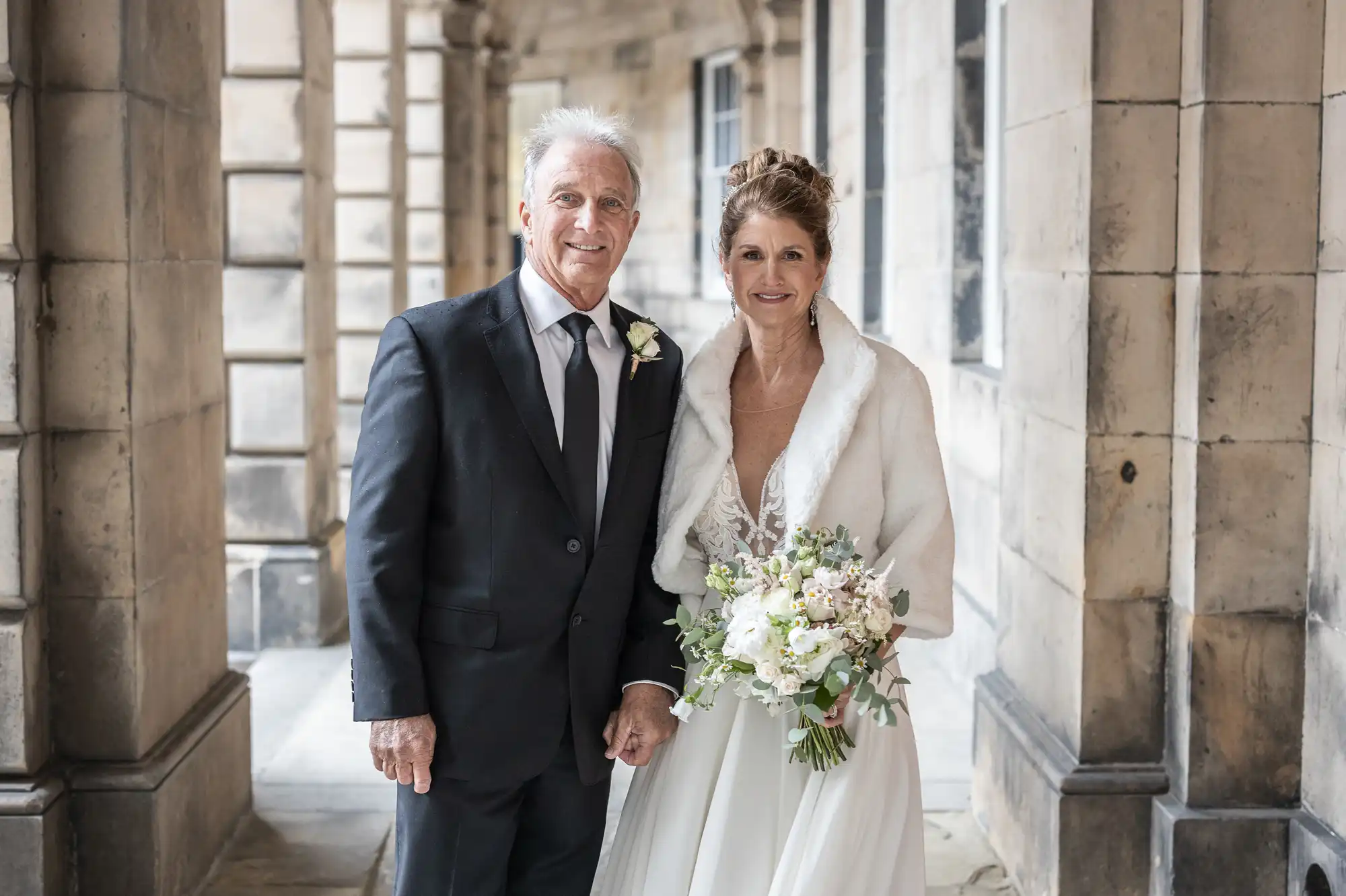 A couple in formal attire stands in a stone corridor. The man wears a dark suit, and the woman wears a white gown with a fur stole and holds a flower bouquet. Both are smiling.