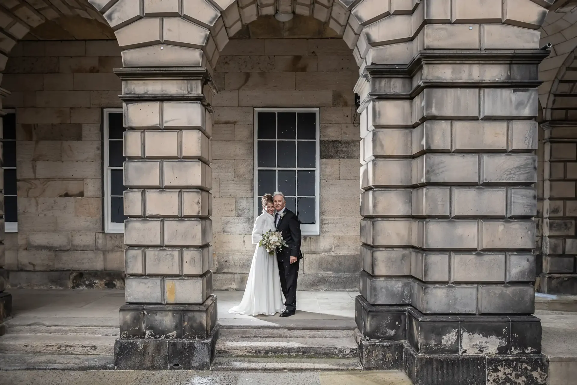Destination wedding photographer: A couple dressed in a wedding attire pose together between stone archways in front of a window on a beige stone building.