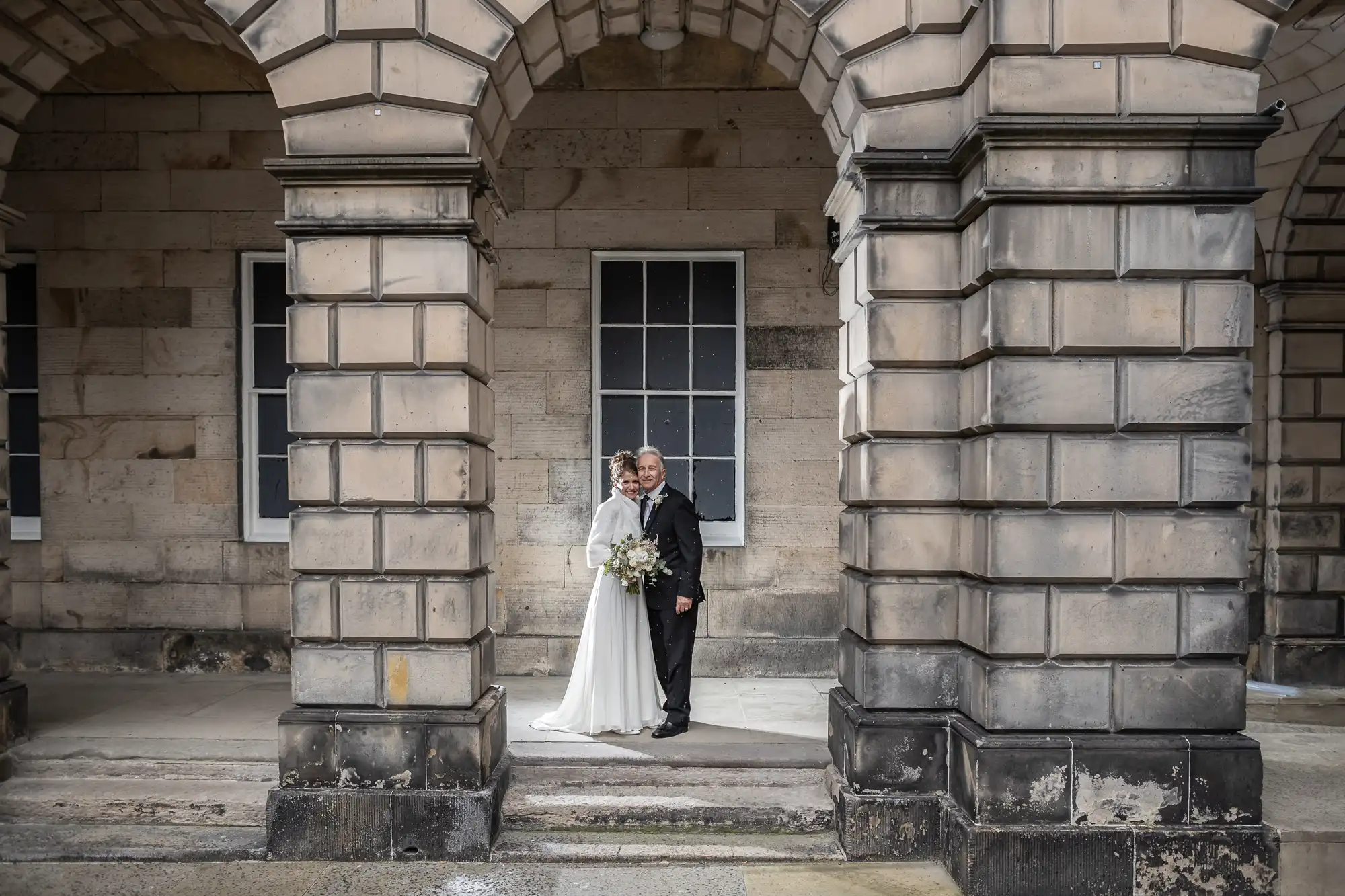 A couple dressed in a wedding attire pose together between stone archways in front of a window on a beige stone building.