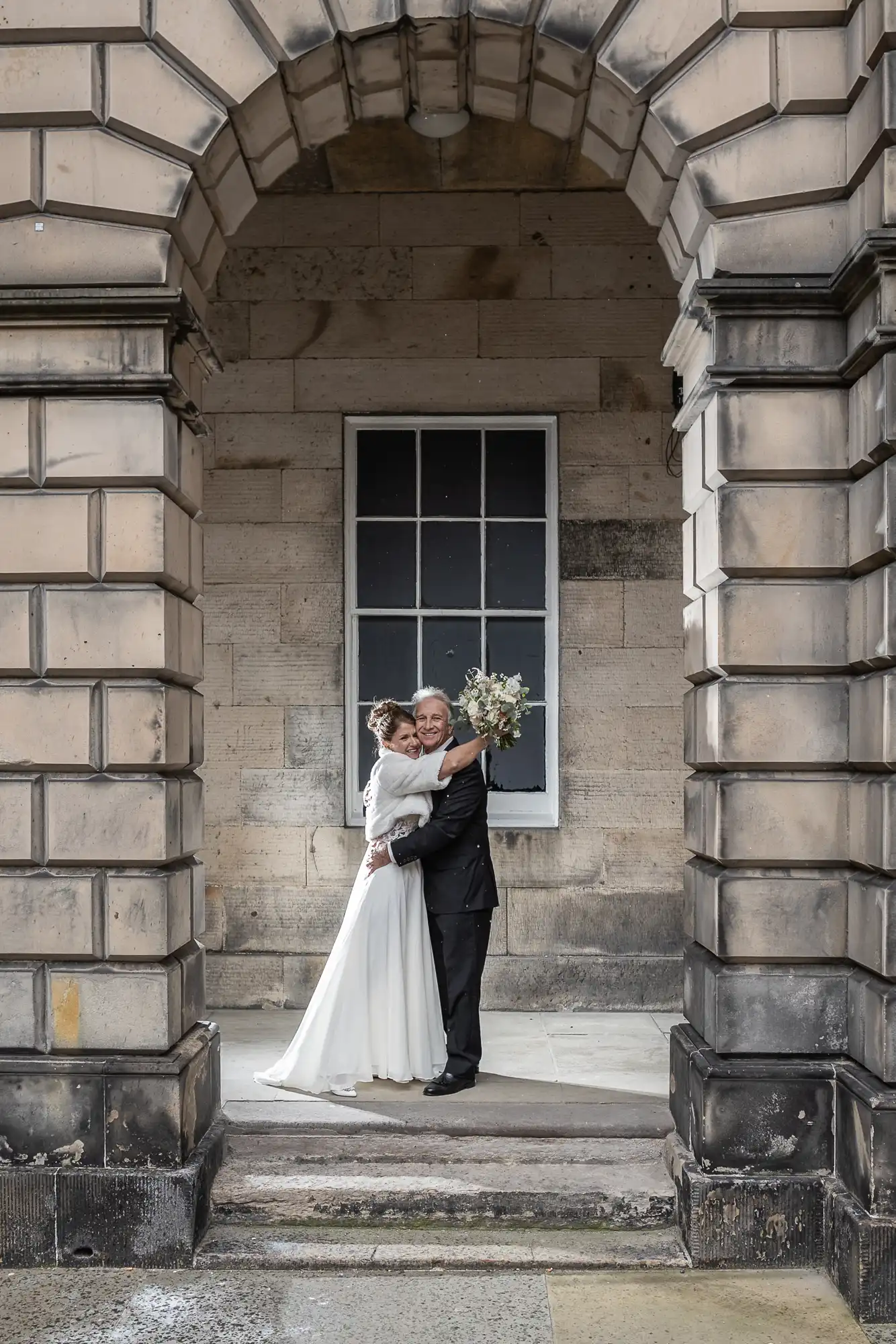 A bride in a white dress and a groom in a black suit stand hugging under a stone archway with a large window in the background. The bride holds a bouquet of flowers.