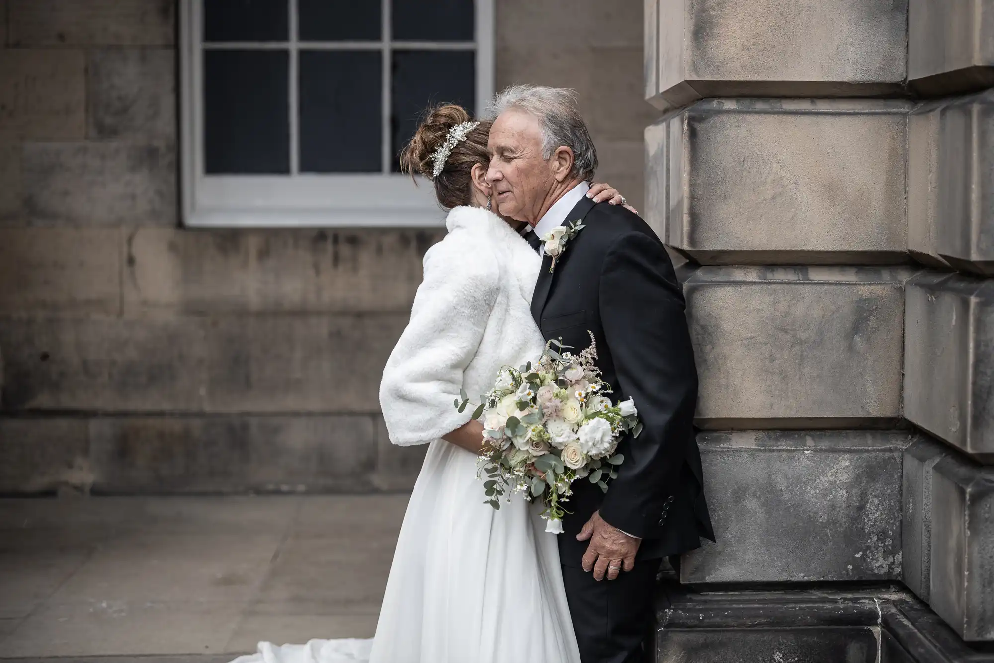 An older man in a suit and boutonniere embraces a woman in a white wedding dress holding a bouquet, standing by a stone building.