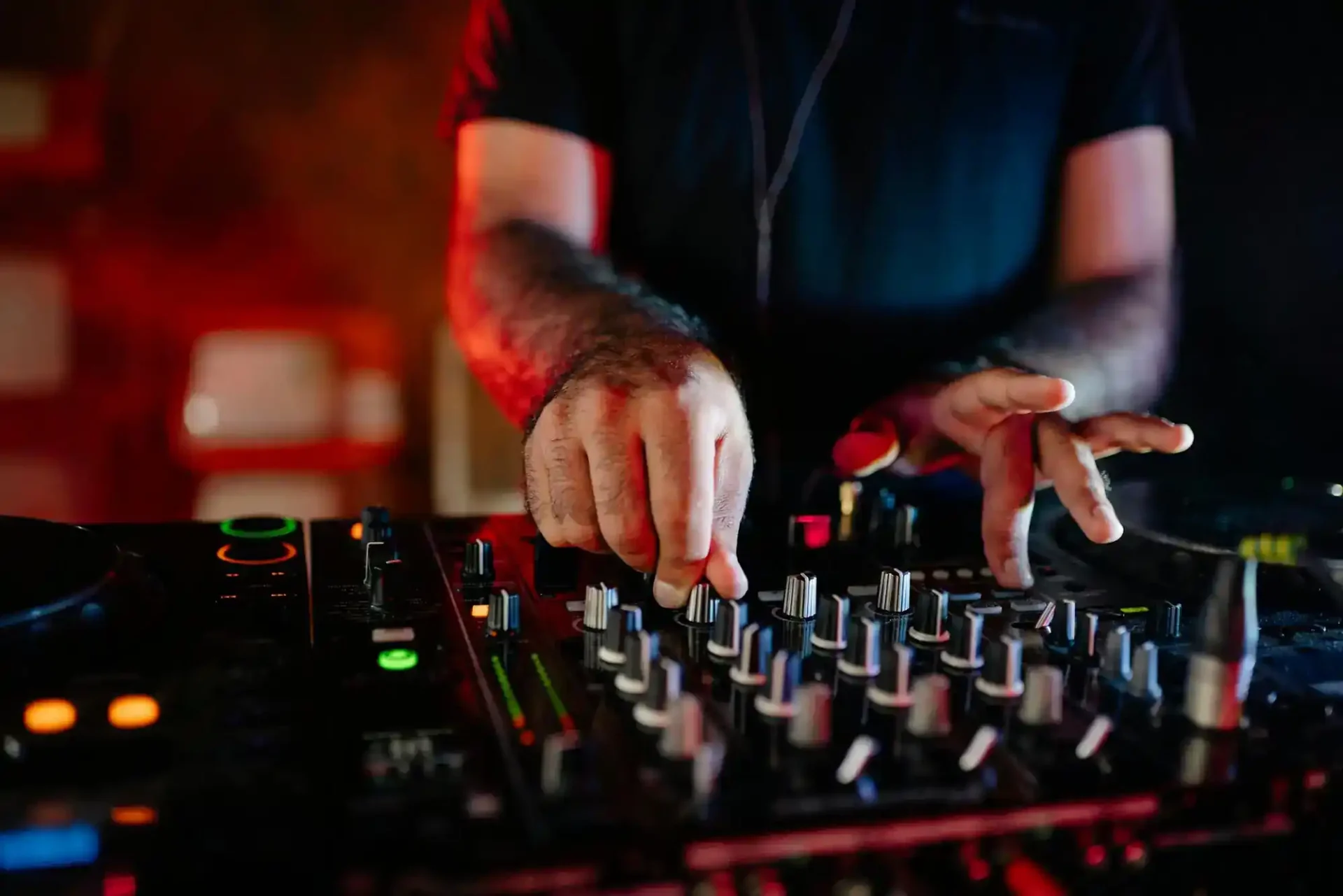 A DJ adjusts knobs and sliders on a mixing desk in a dimly lit setting. Only the DJ’s hands and forearms are visible, focusing on the equipment. The background is blurred with red and orange lighting, emphasising a nightclub or party atmosphere.