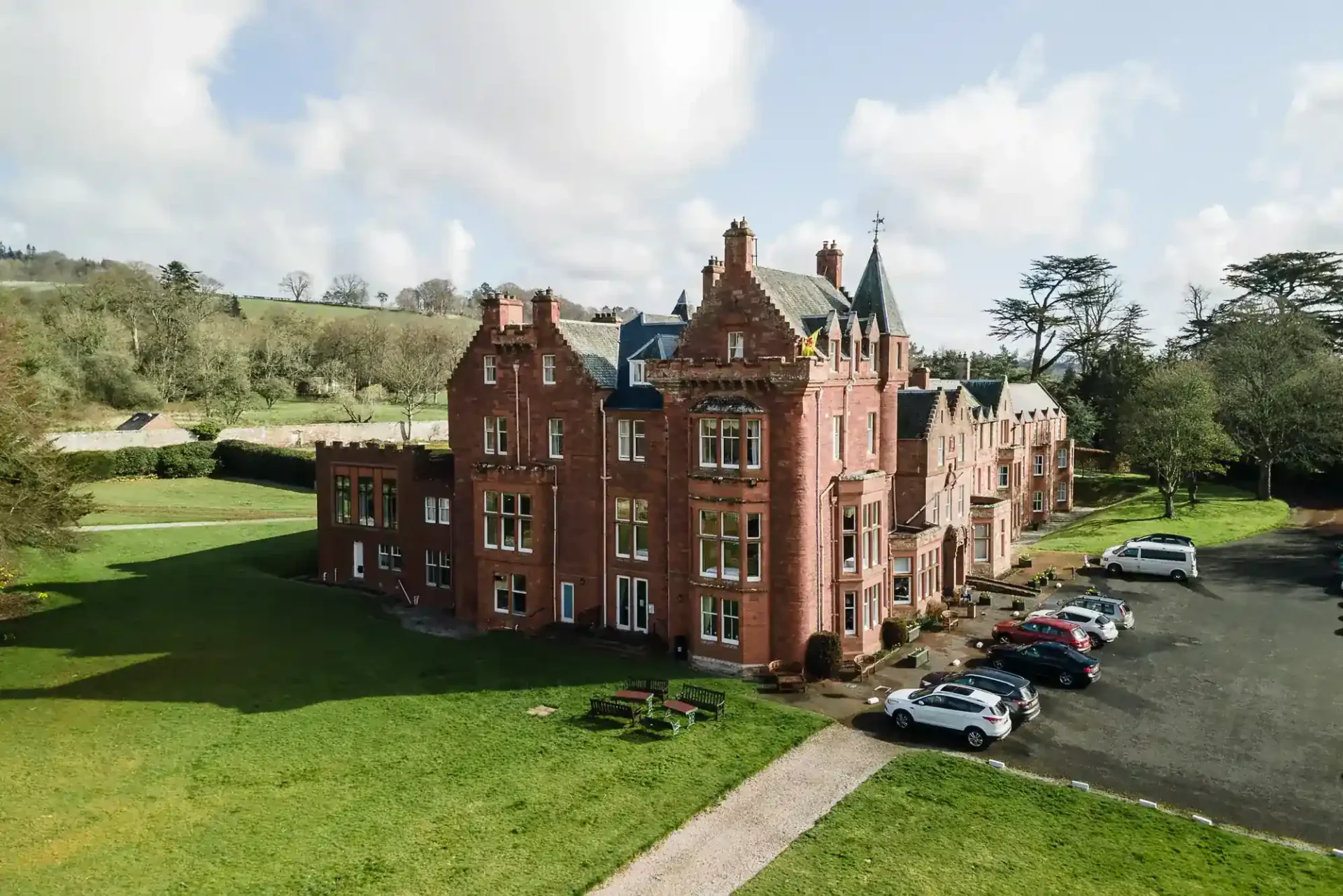 A large red-brick Victorian mansion, Dryburgh Abbey Hotel, with gabled roofs and turrets sits on a green lawn. Several cars are parked in a car park to the right, and rolling hills with trees are visible in the background under a partly cloudy sky.