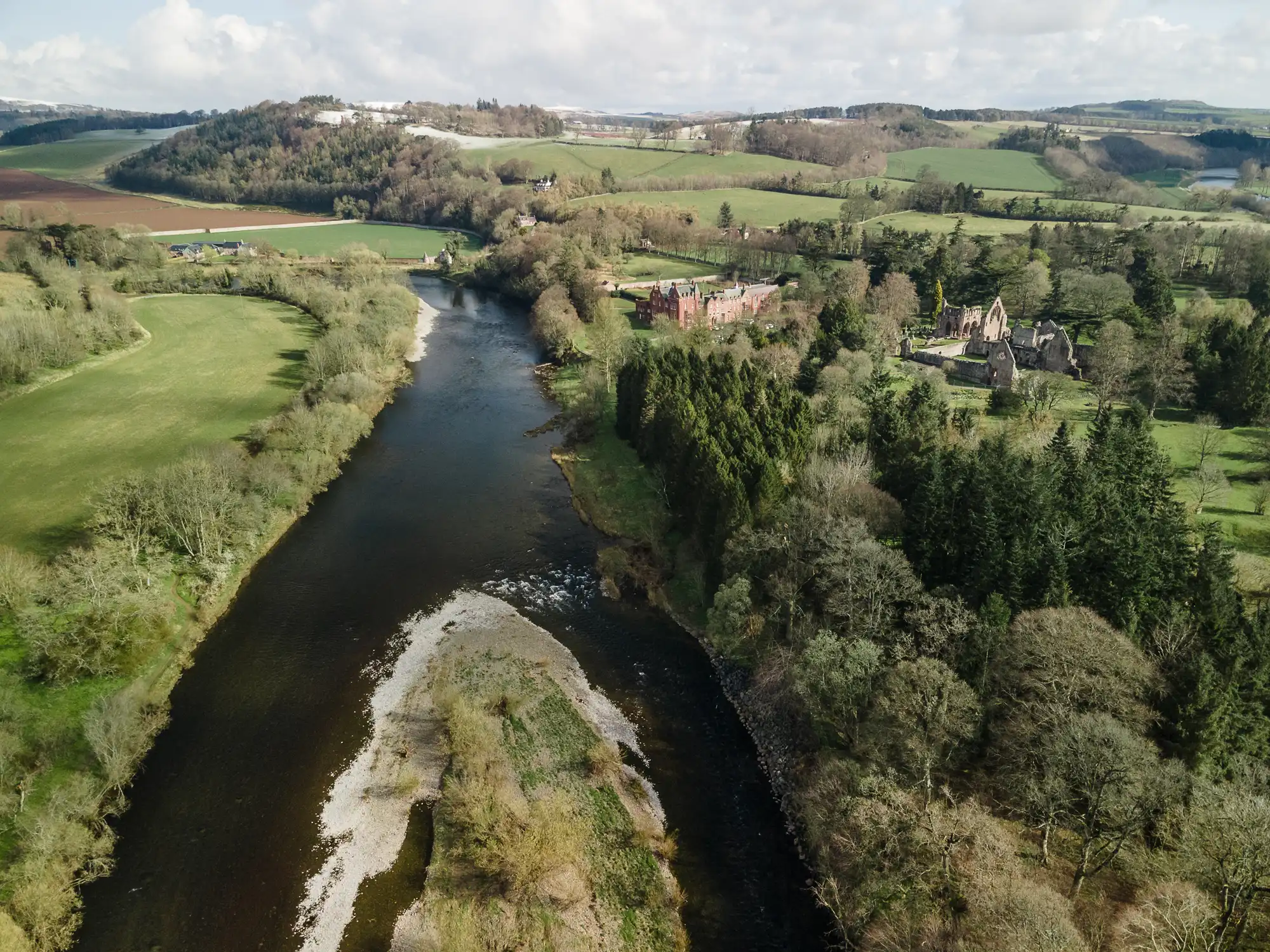 A wide river winds through a lush, green countryside with open fields, dense trees, and rolling hills. On the right, a large stone building and a castle-like structure sit among the trees. The sky is partly cloudy, casting soft light over the landscape.