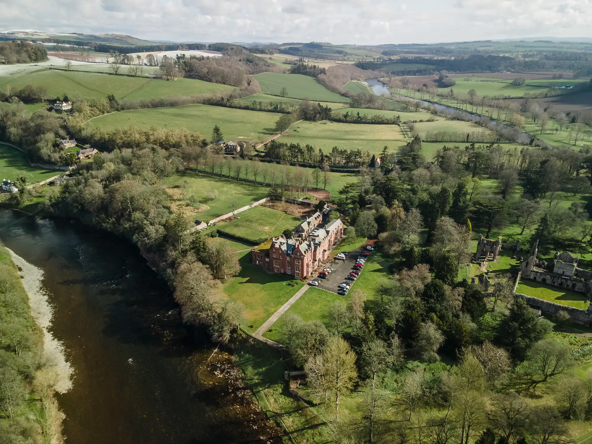 A red-brick mansion sits beside a river, surrounded by trees and expansive green fields. Multiple cars are parked outside the building. Rolling hills, smaller houses, and patches of forest extend into the distance under a partly cloudy sky.