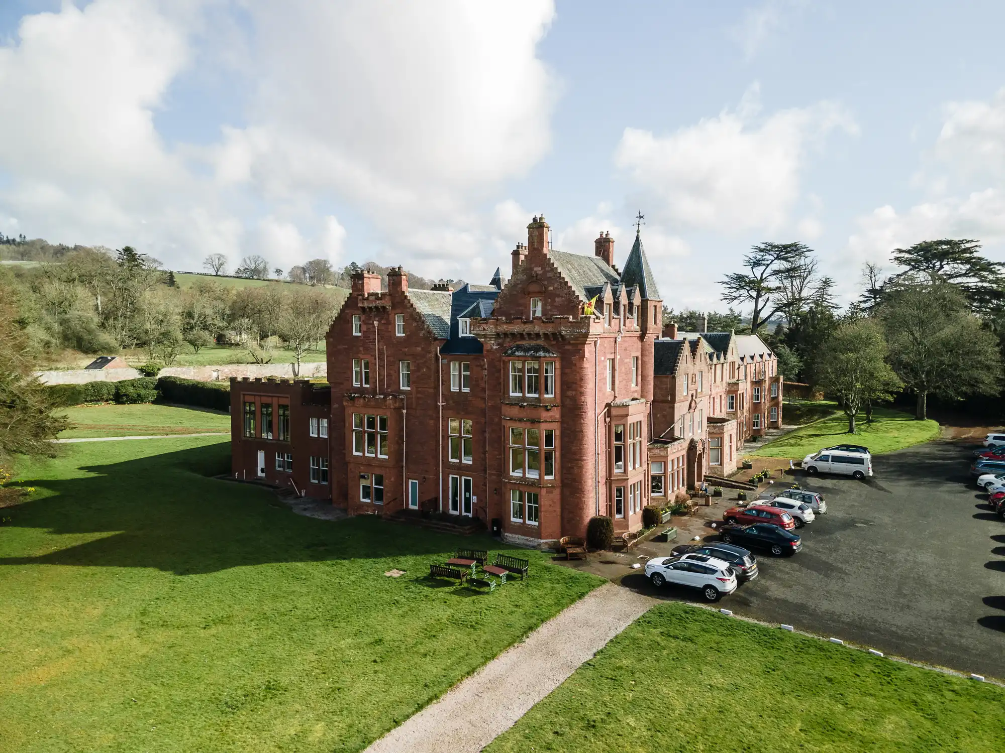 A large, red-brick Victorian mansion with ornate chimneys sits on a lush green lawn. Numerous parked cars line a gravel car park to the right. Trees and rolling hills surround the building under a bright, partly cloudy sky.