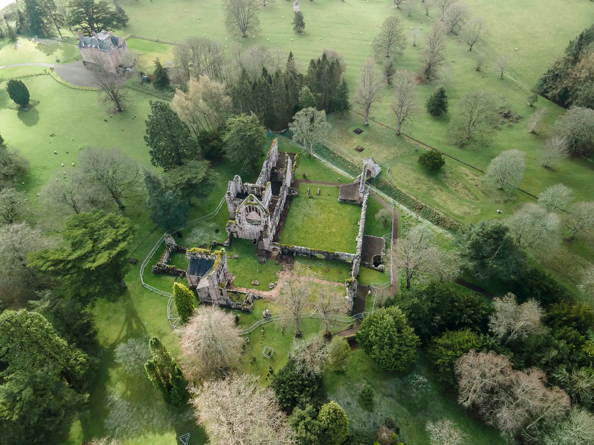Aerial view of an old stone abbey ruin surrounded by a rectangle of green grass and trees. Ruined walls and arches remain, with roofless sections. Nearby is a smaller building; open countryside and scattered trees surround the site.