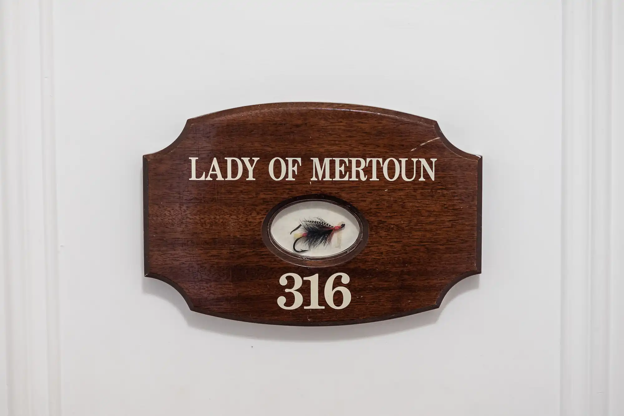 A wooden plaque on a white door reads "LADY OF MERTOUN" in white letters. Below, a small display holds a decorative fishing fly, and the number "316" is printed at the bottom of the plaque.
