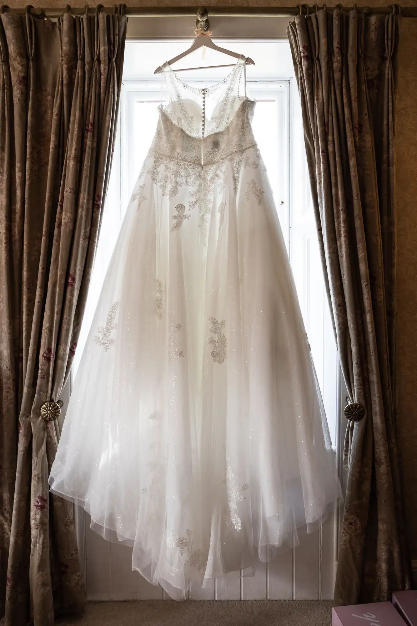 A white, sleeveless wedding dress with a full, flowing tulle skirt and delicate lace appliqué details hangs on a hanger in front of a sunlit window, framed by dark patterned curtains tied back on each side.