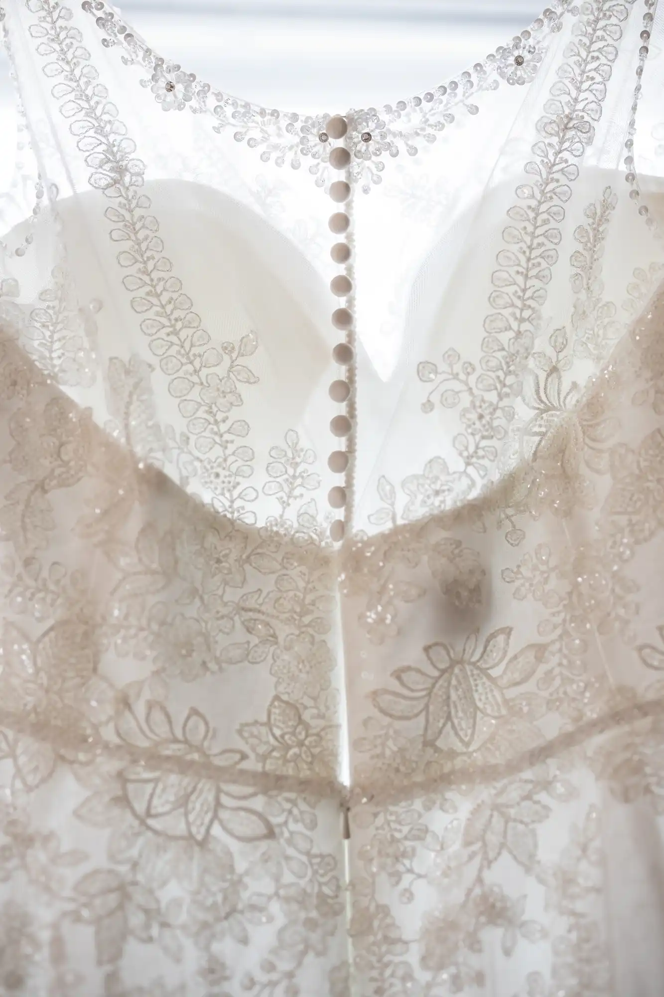 Close-up of the back of a white wedding dress featuring sheer fabric, delicate floral lace embroidery, and a row of small round buttons running down the centre. Light shines softly through the translucent material, highlighting the intricate pattern.