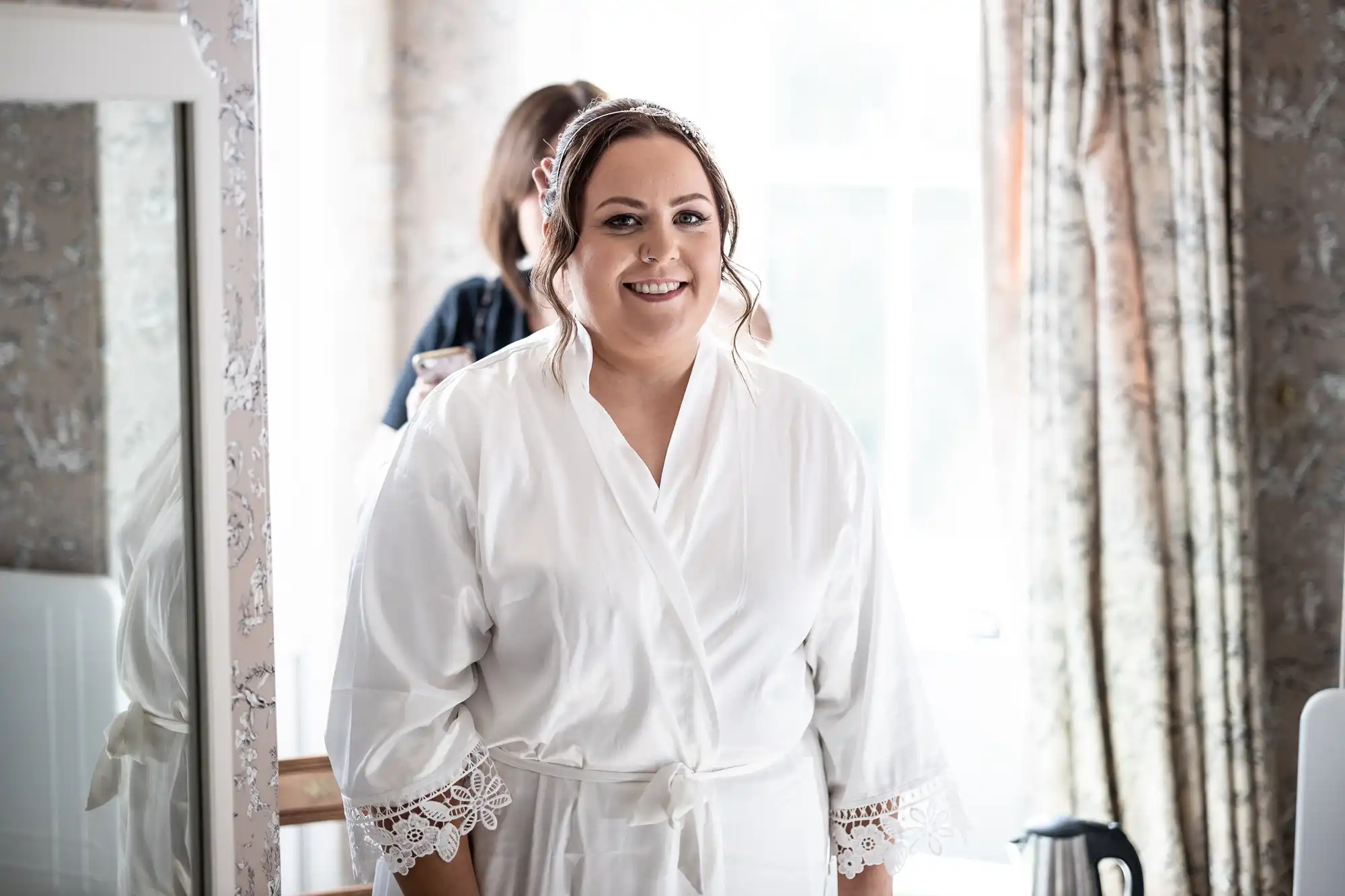 A smiling woman in a white dressing gown with lace-trimmed sleeves stands indoors. Her brown hair is styled, and she wears a hairband. Light from a window brightens the room, and floral curtains and a mirror are visible in the background.
