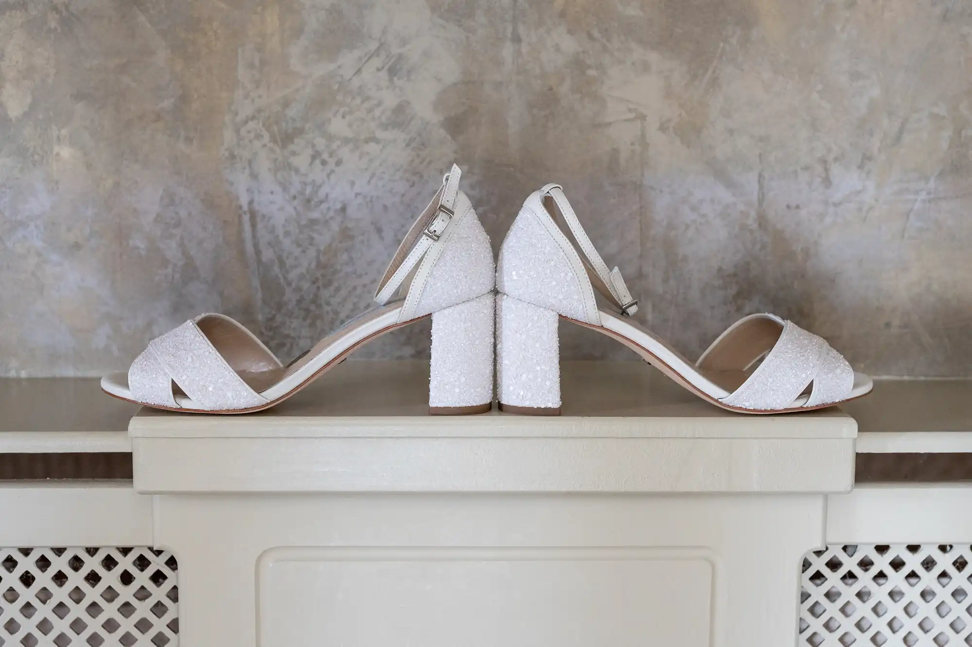 Two sparkly white heeled sandals are placed back to back on a cream-coloured ledge. Each sandal features an ankle strap with a buckle and a closed block heel, set against a textured grey and beige wall.