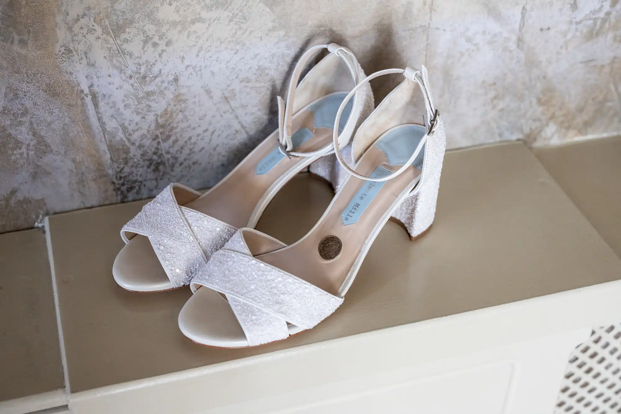 A pair of white, glittery, open-toed block-heeled sandals with ankle straps is placed side by side on a beige surface against a textured, light-coloured wall. The shoes have criss-cross front straps and a light blue insole.