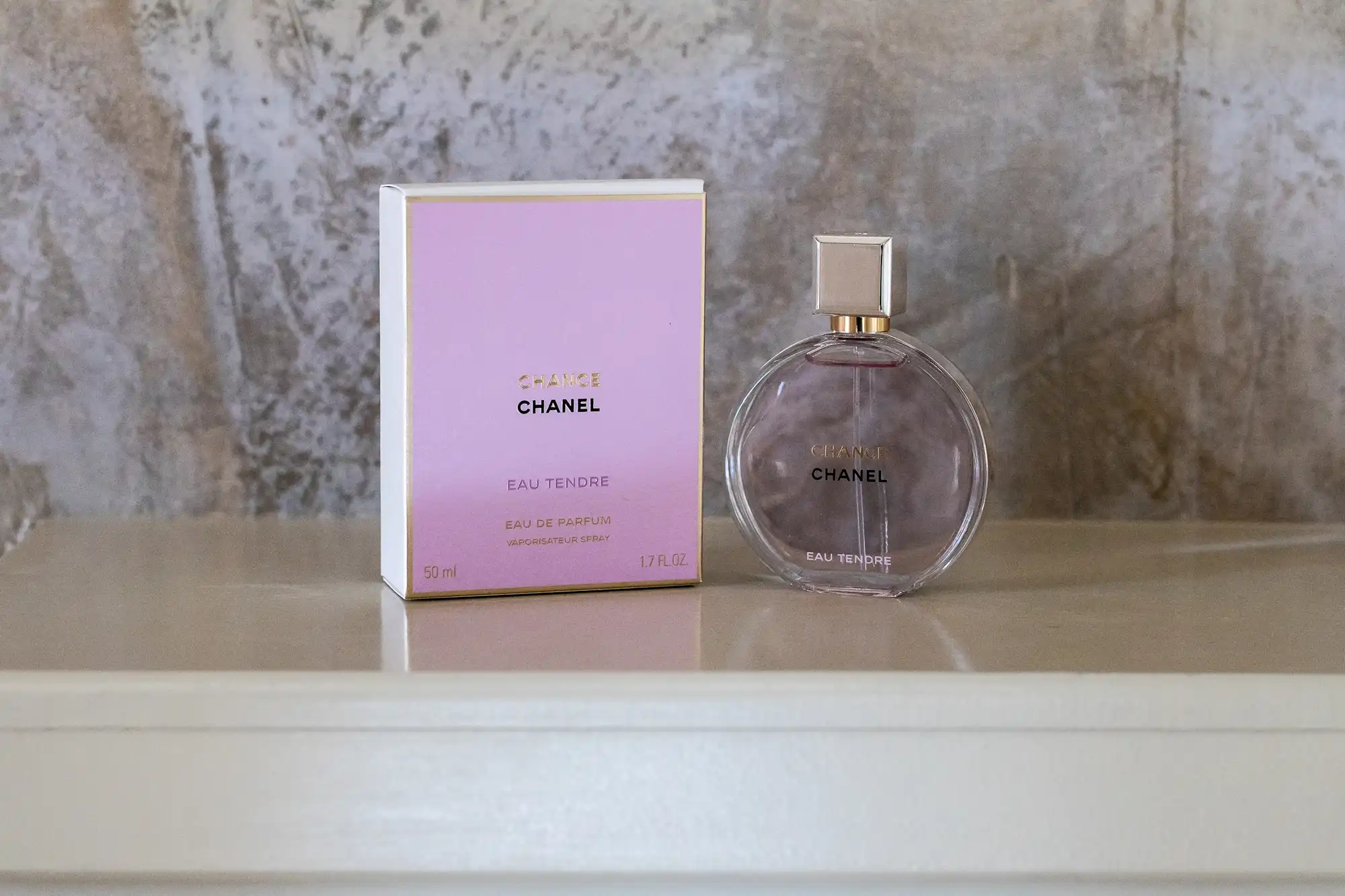 A round glass bottle of Chanel Chance Eau Tendre perfume stands next to its pale pink and gold box on a cream-coloured surface, with a marbled beige and grey background.