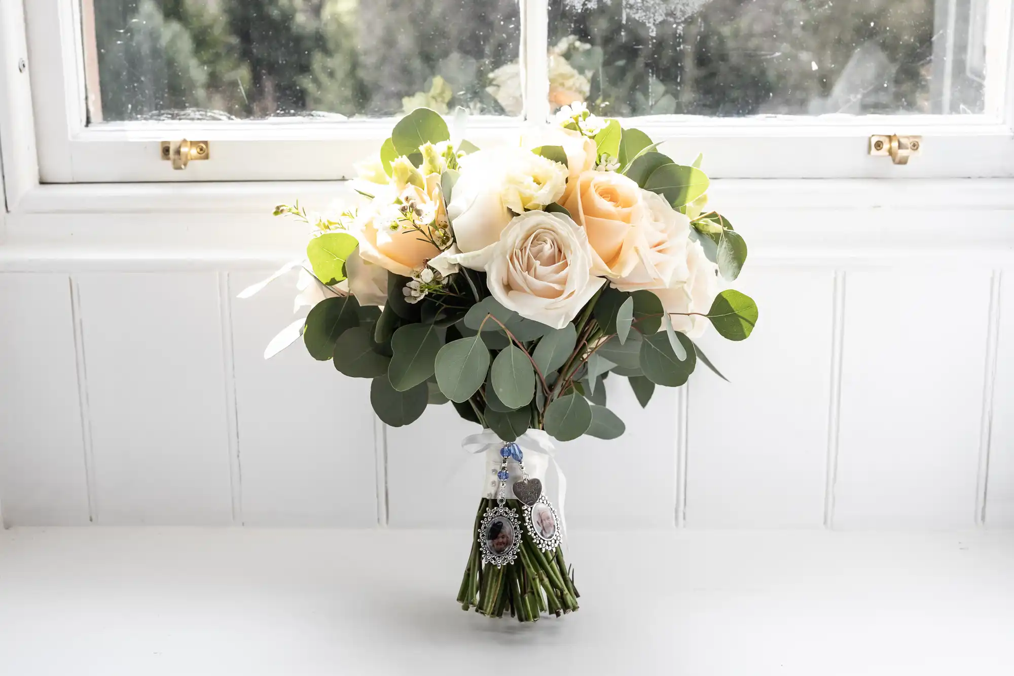 A bouquet of cream and blush roses mixed with eucalyptus leaves sits on a white windowsill. The bouquet is tied with white ribbon and decorated with two small silver photo charms. Sunlight streams through the window, softly illuminating the flowers.